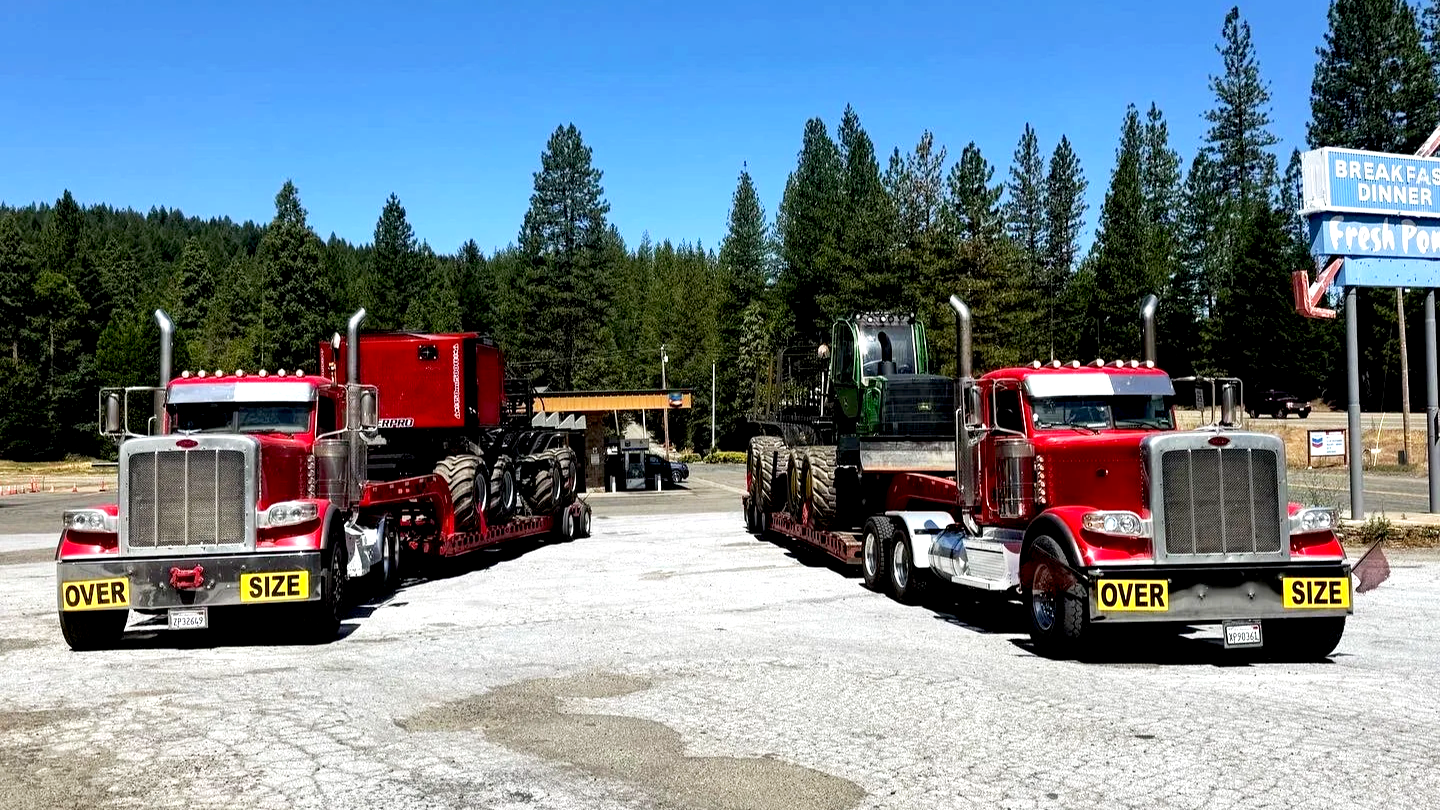 Two large red trucks from H&S Heavy Haul carrying oversized vehicles, one with a green tractor and the other with a red tractor, parked in an outdoor lot with a sign showing breakfast and dinner options and a forested background.