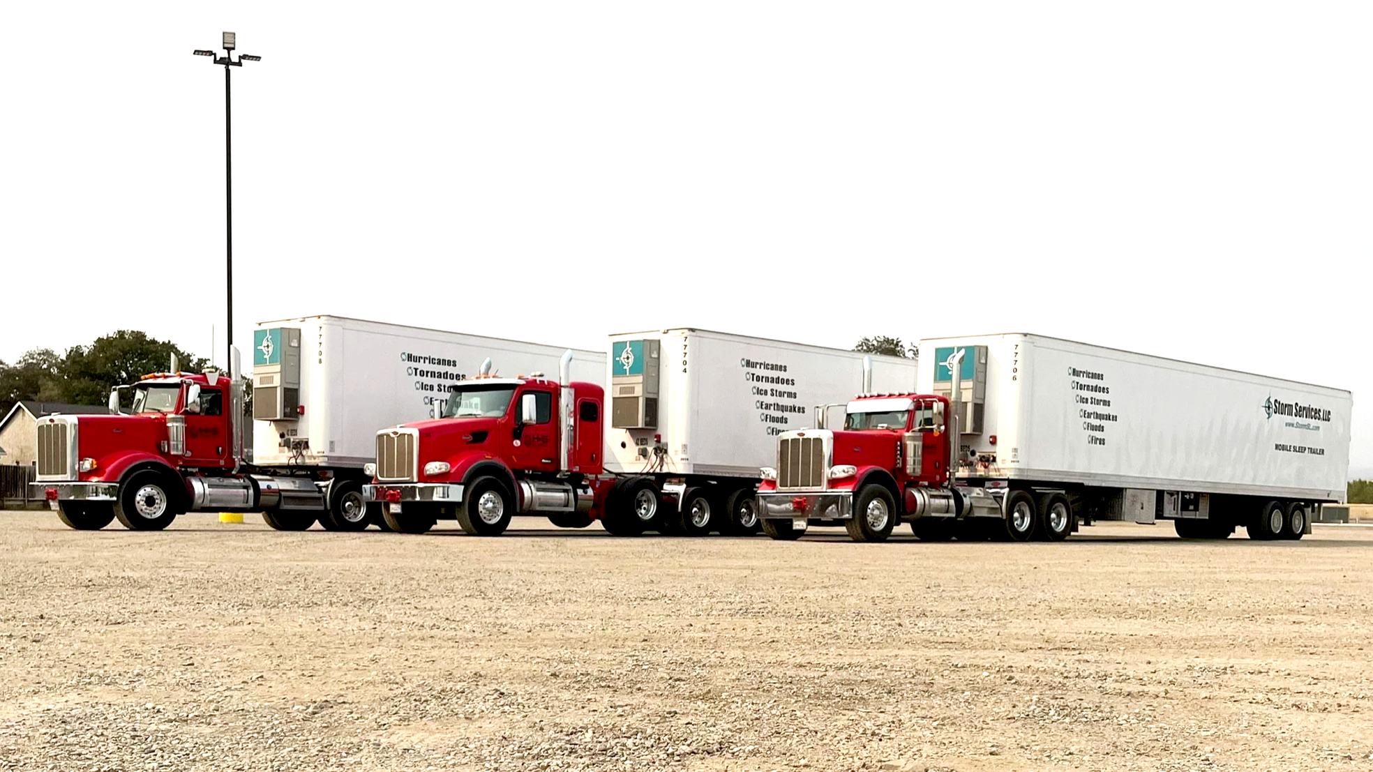 Three red semi-trucks from H&S Heavy Haul with white trailers parked on a gravel lot, with a pole and some trees in the background.