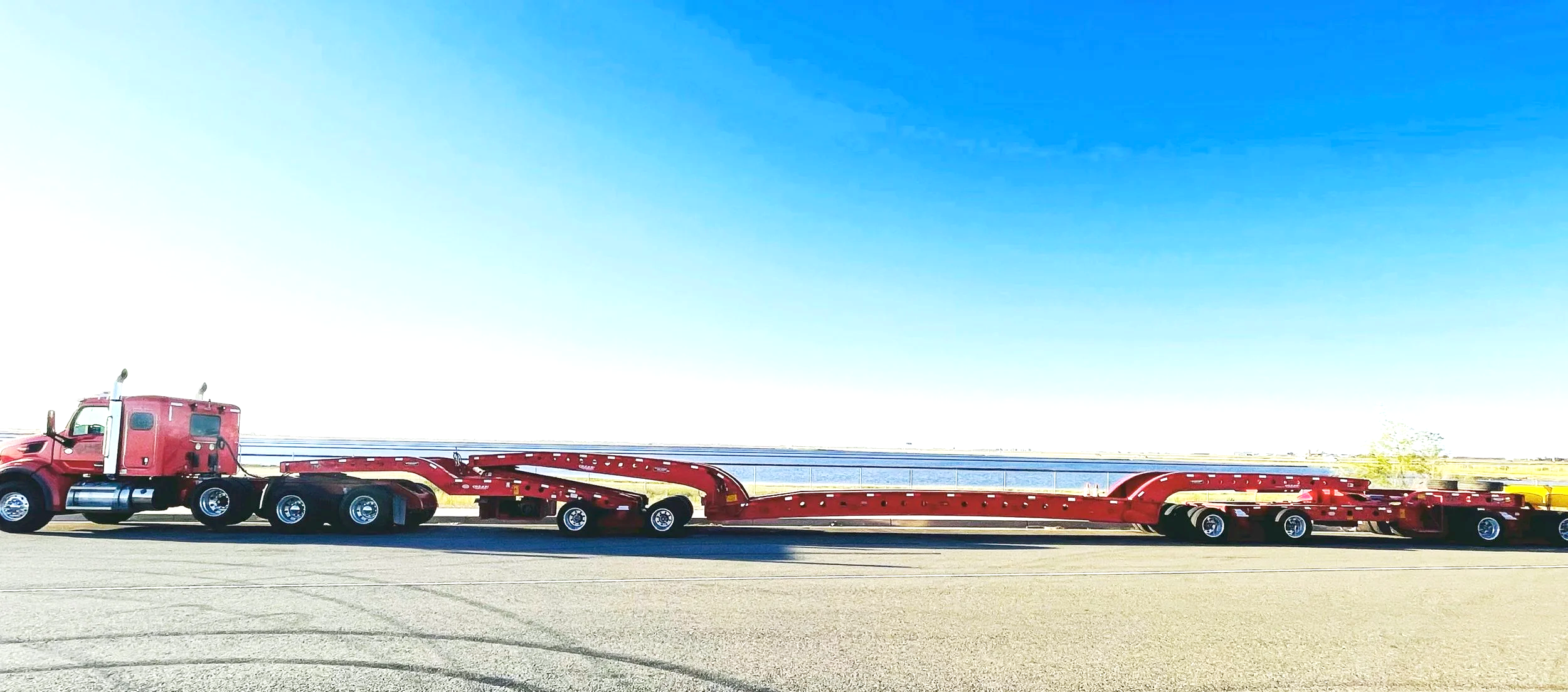 Red semi-truck from H&S Heavy Haul with a flatbed trailer parked on an empty road next to a body of water under a clear blue sky.