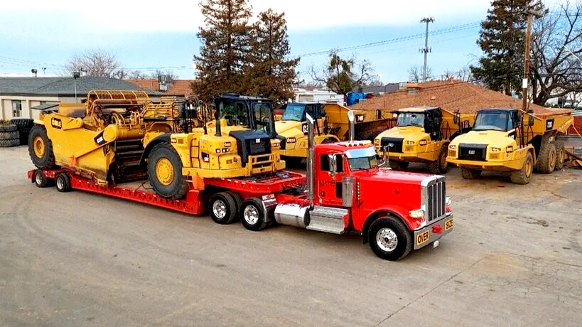 Red semi-truck from H&S Heavy Haul transporting construction machinery, including bulldozers and loaders, parked on a gravel lot with trees and buildings in the background.