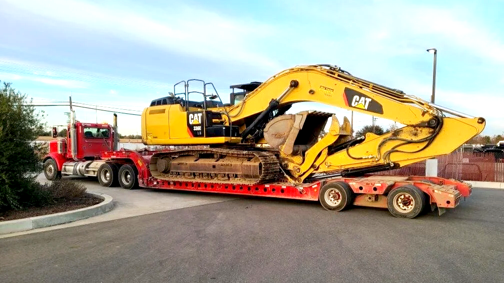 A red semi-truck from H&S Heavy Haul carrying a large yellow CAT excavator on its flatbed trailer.