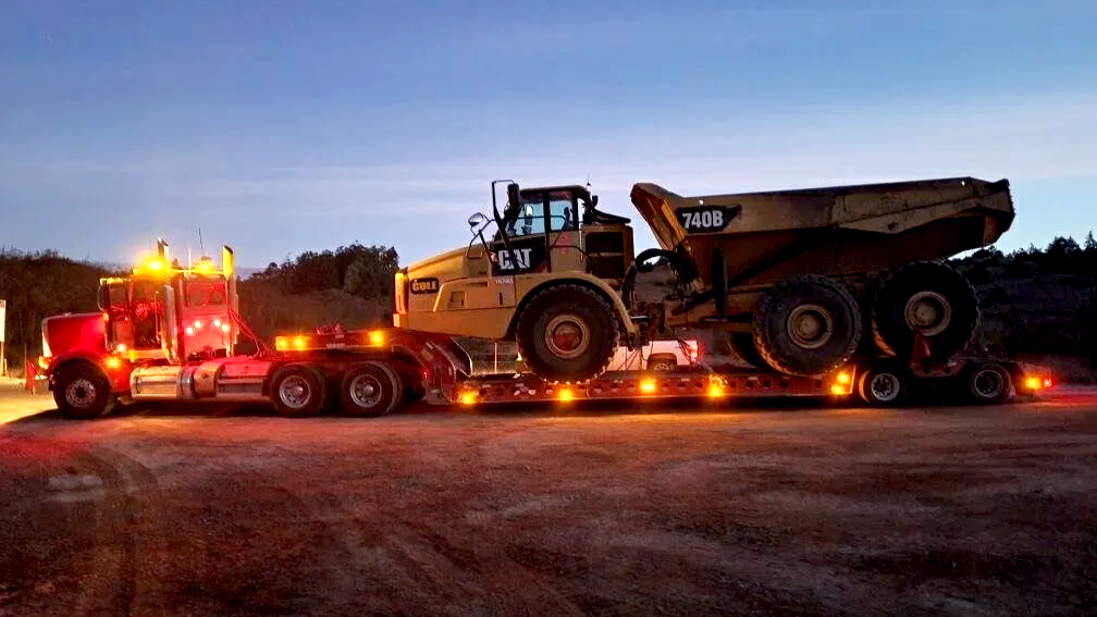 A large semi-truck from H&S Heavy Haul transporting a CAT 740B articulated dump truck during dusk or dawn, with lights on the semi-truck illuminating the scene.