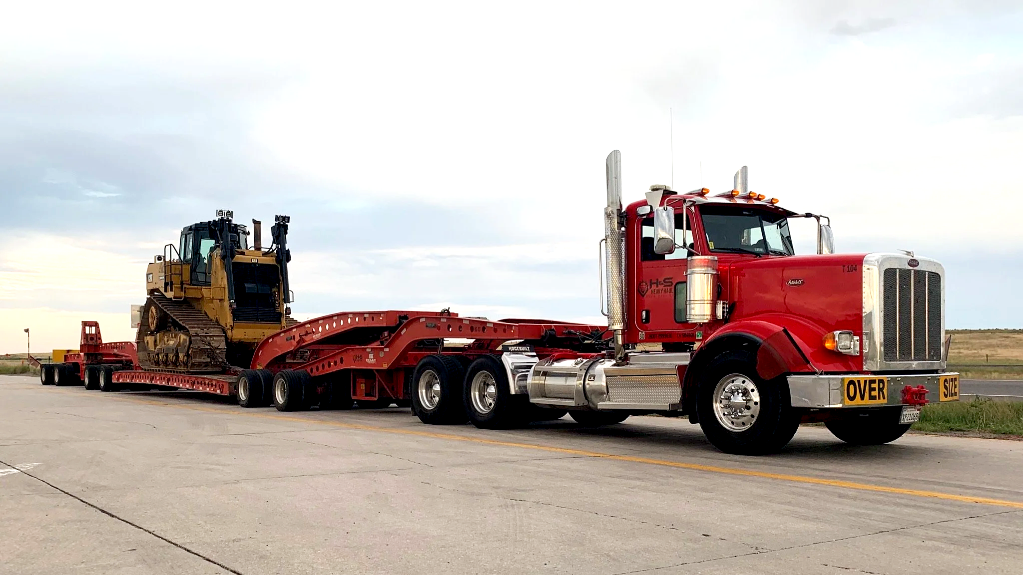 A red semi-truck from H&S Heavy Haul transporting a large bulldozer on a flatbed trailer on an empty road with a cloudy sky in the background.