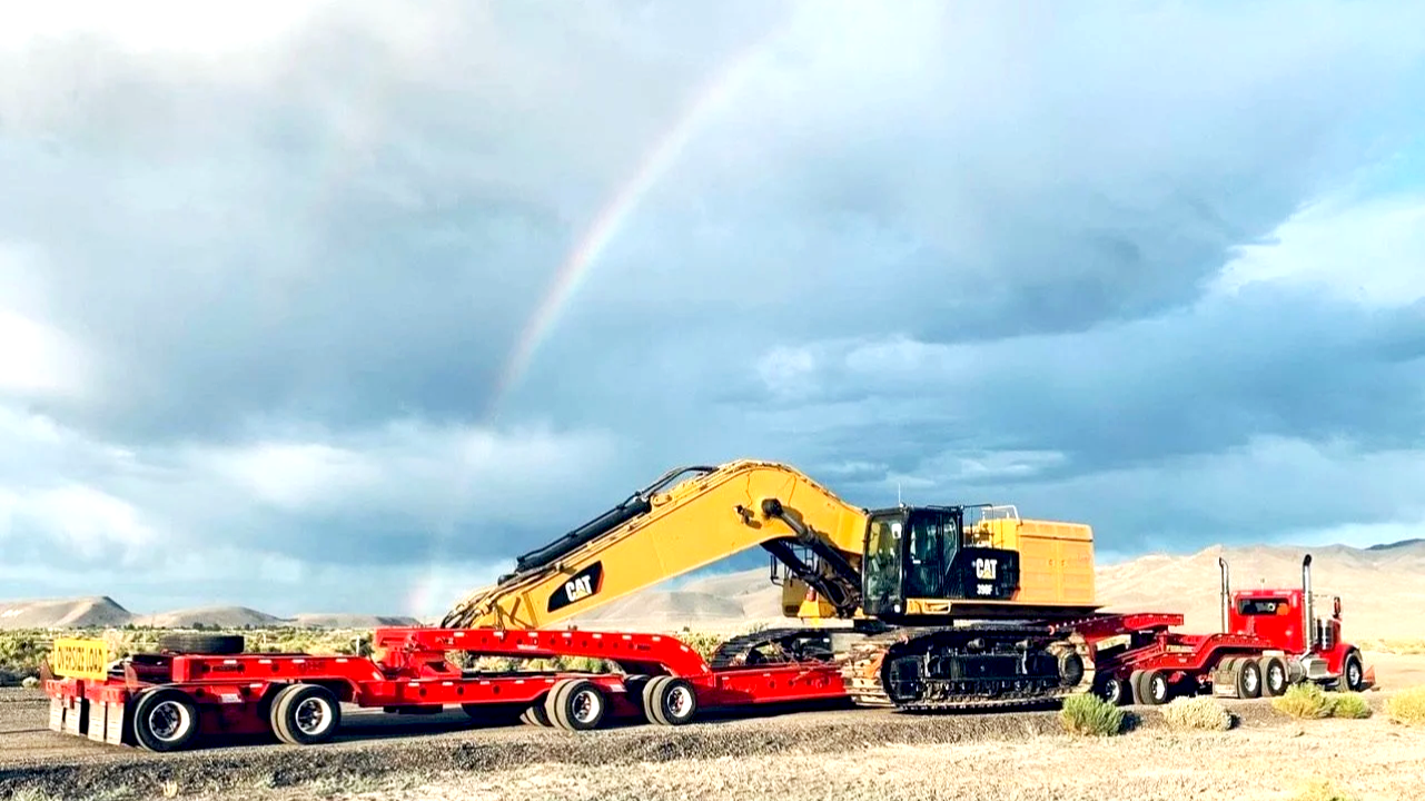 A yellow Caterpillar excavator on a red flatbed truck from H&S Heavy Haul in a desert landscape with hills and a rainbow in the sky.