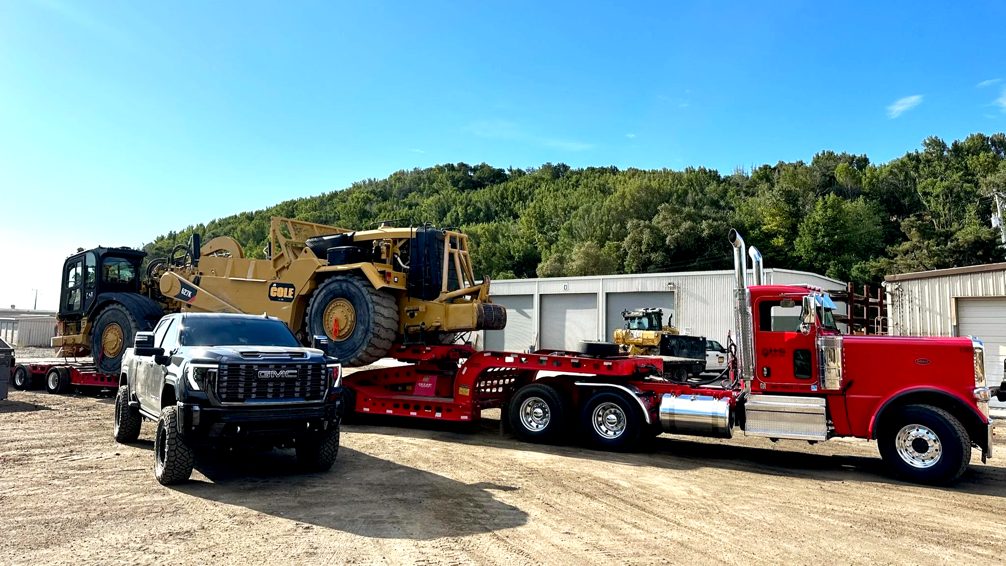 A red semi-truck from H&S Heavy Haul with a flatbed trailer carrying construction equipment, including a large yellow bulldozer, parked outside a warehouse with a forested hillside in the background.