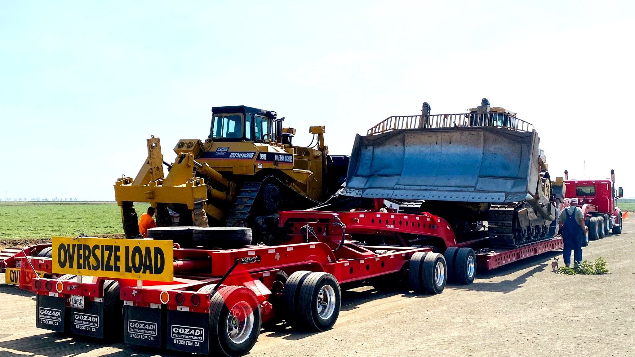 A large red semi-truck from H&S Heavy Haul with a trailer carrying heavy machinery, including a bulldozer. A yellow sign on the trailer reads 'OVERSIZE LOAD'. People are standing around.