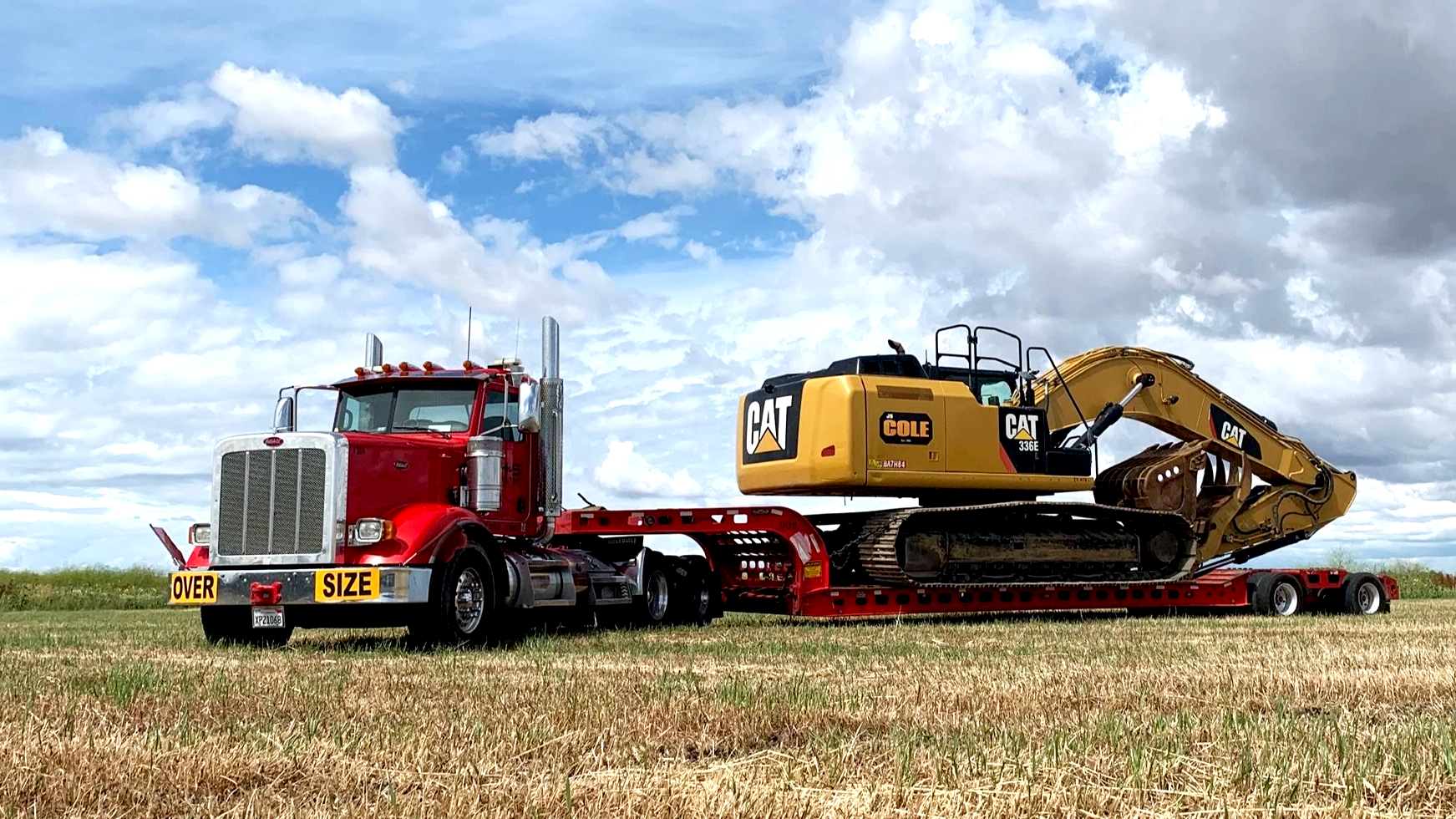 A red H&S Heavy Haul semi-truck with a flatbed trailer carrying a yellow CAT excavator, set against a partly cloudy sky in an open field.