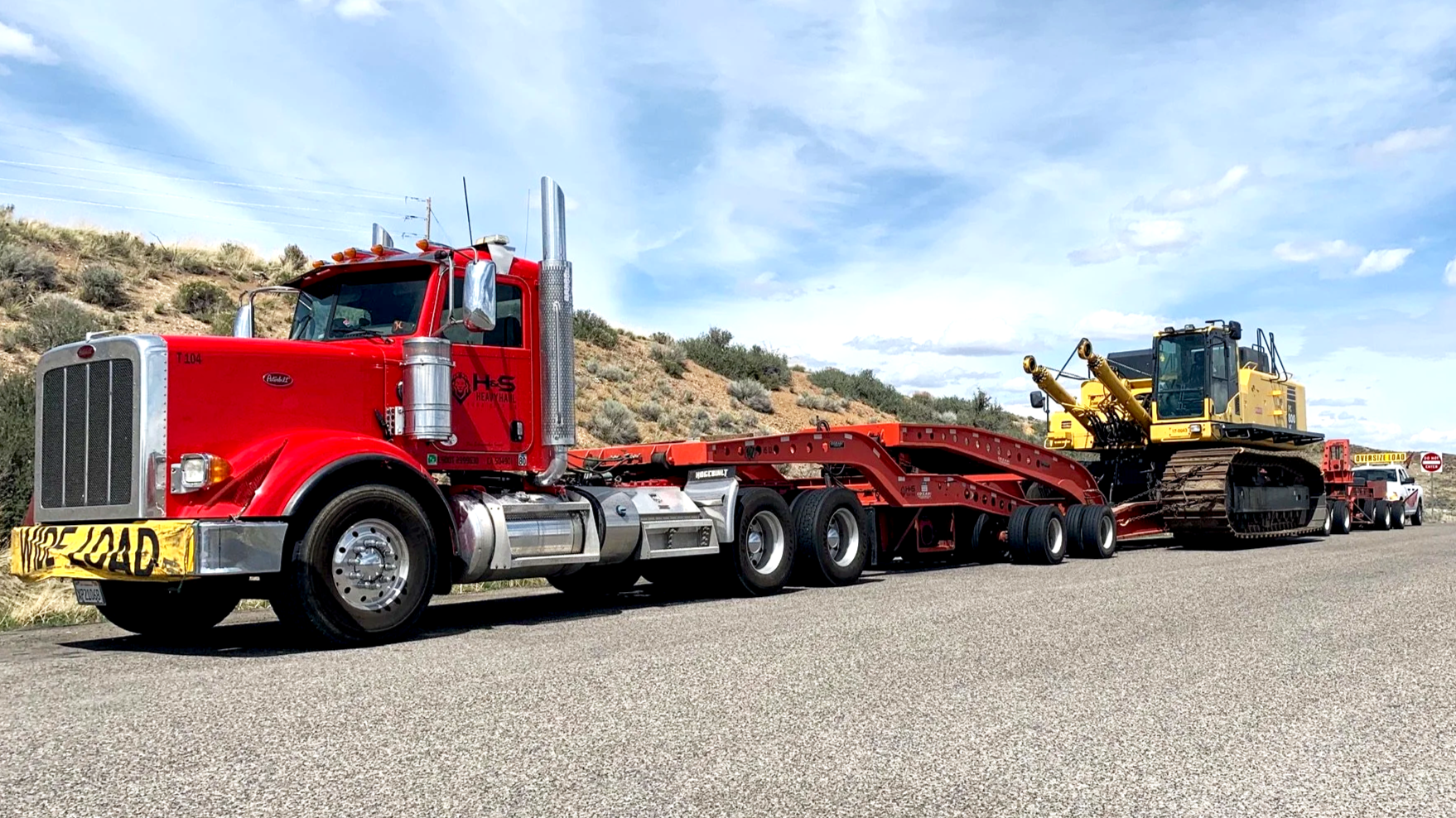 A red heavy-duty semi-truck from H&S Heavy Haul with a flatbed trailer transporting a large yellow bulldozer on a highway near a hilly area with sparse vegetation. The sky is partly cloudy.