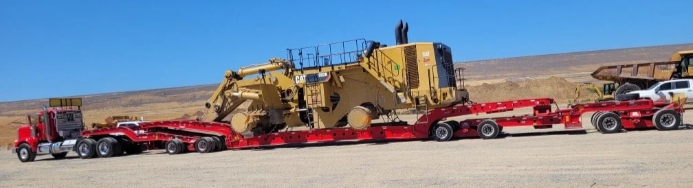 A large yellow Caterpillar (CAT) excavator loaded on a red flatbed trailer from H&S Heavy Haul, parked on a dirt or gravel road with a partly cloudy sky and barren landscape in the background.