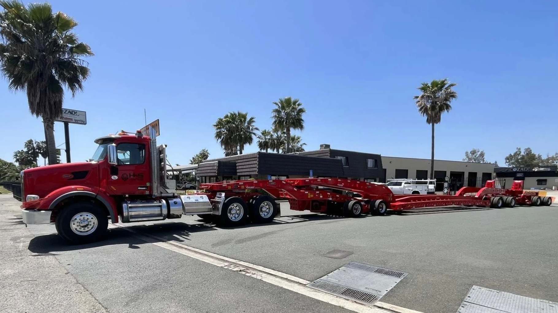 A red H&S Heavy Haul truck and trailer is parked on a street with palm trees and buildings in the background under a clear blue sky.