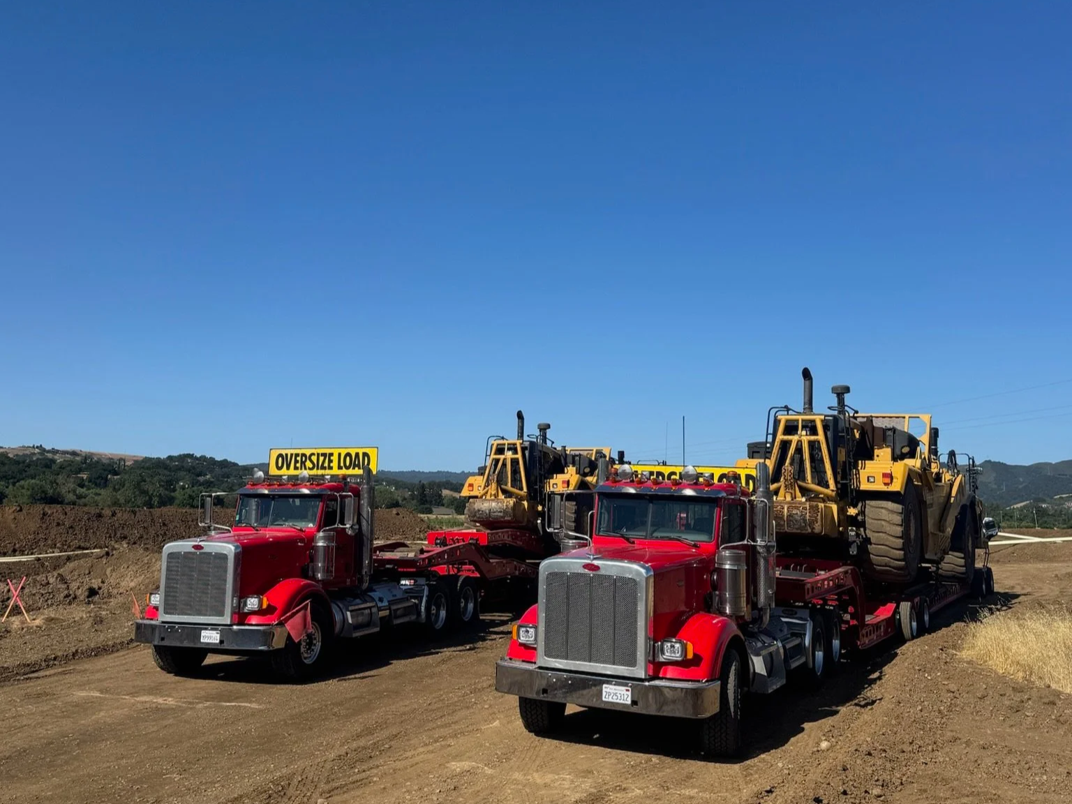 Two red flatbed trucks from H&S Heavy Haul carrying yellow construction bulldozers on a construction site with dirt and hills in the background under a clear blue sky.