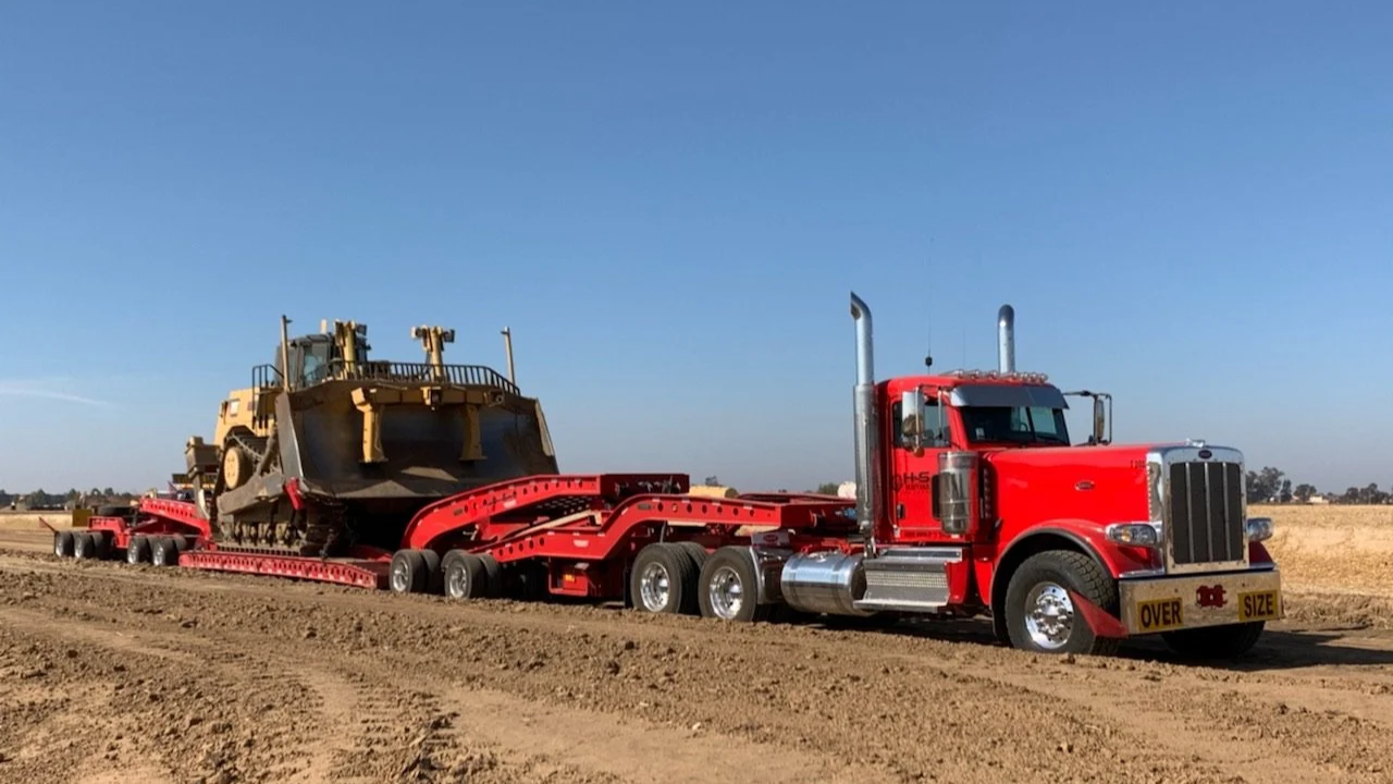 A red semi-truck from H&S Heavy Haul with a flatbed trailer transporting a large bulldozer across a dirt field under a clear blue sky.