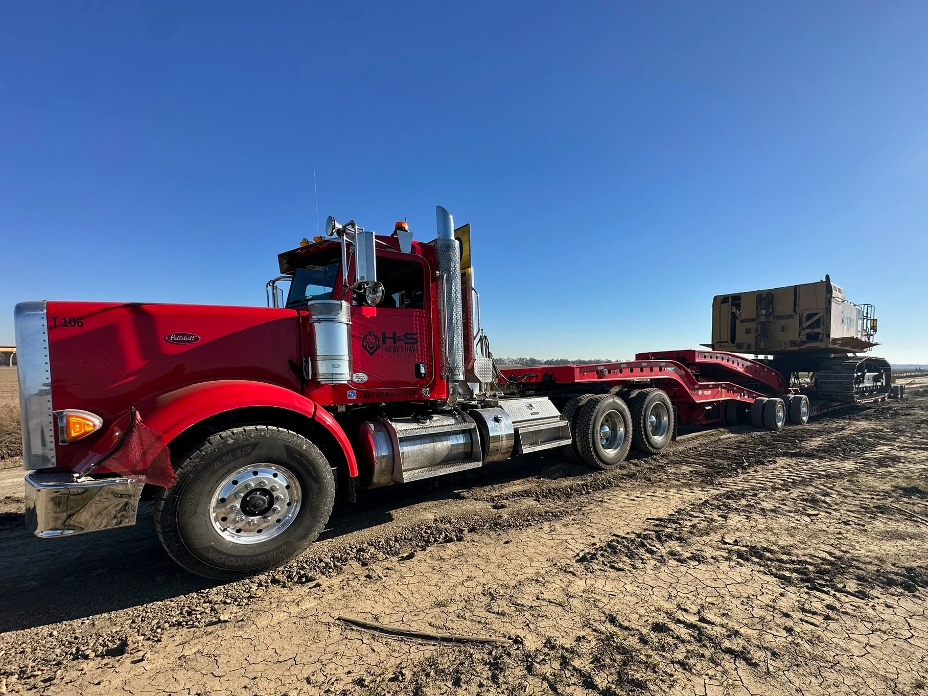 A red heavy-duty truck from H&S Heavy Haul with a flatbed trailer in a dirt field under a clear blue sky.