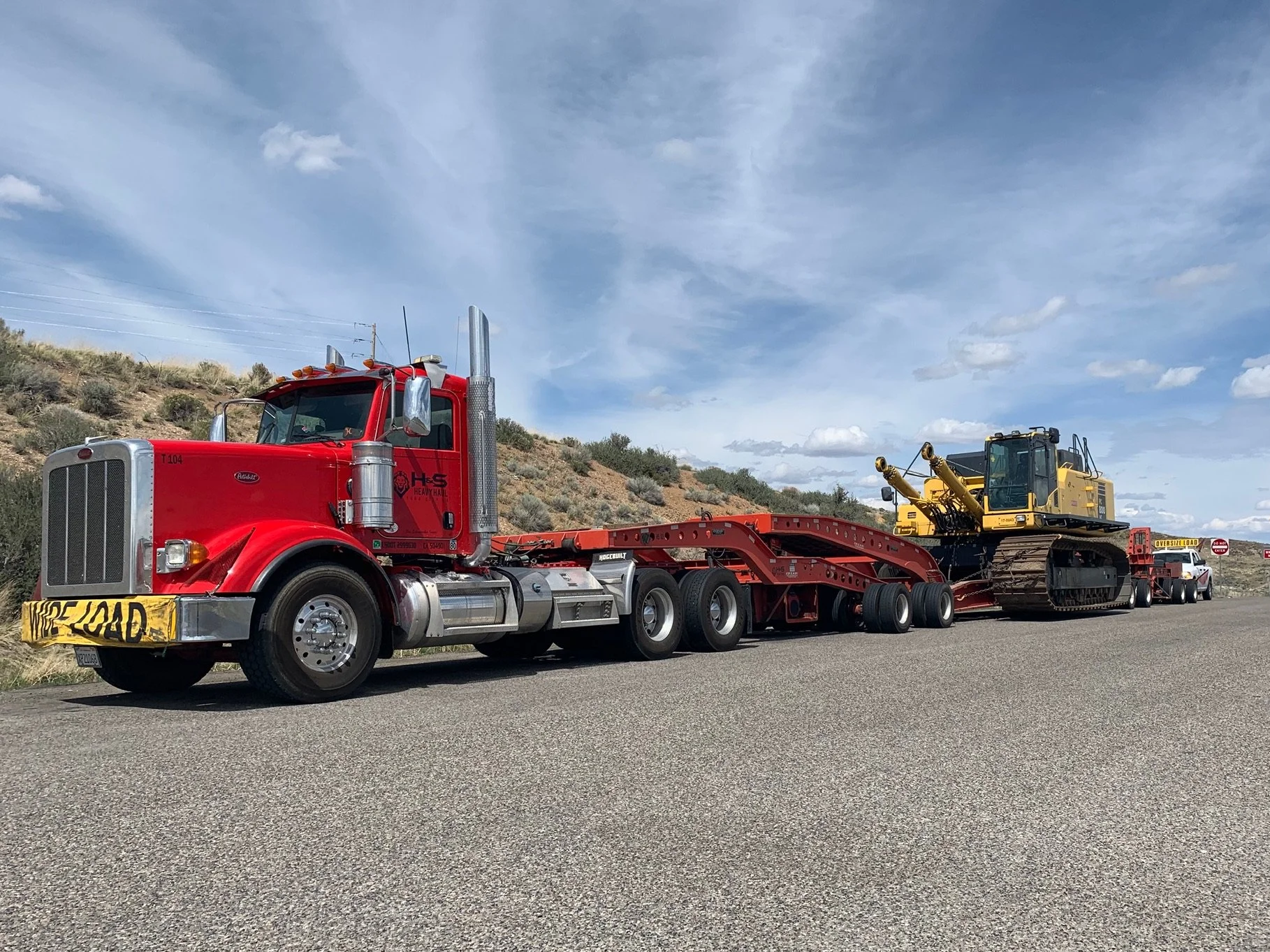 A large red semi-truck from H&S Heavy Haul with a flatbed trailer and a Caterpillar bulldozer being transported on a highway against a backdrop of a hill and a partly cloudy sky.