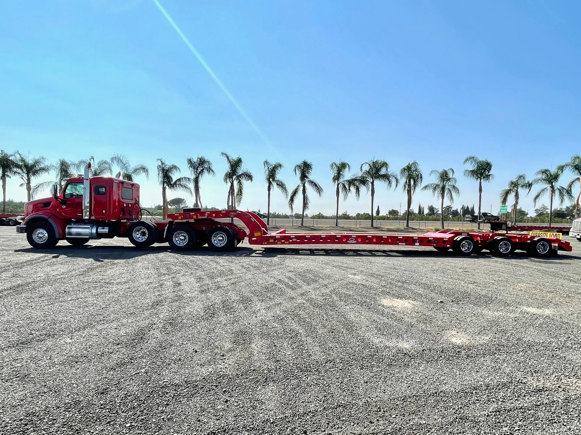 A red semi-truck from H&S Heavy Haul with a flatbed trailer parked on a gravel lot with palm trees in the background and a clear blue sky.