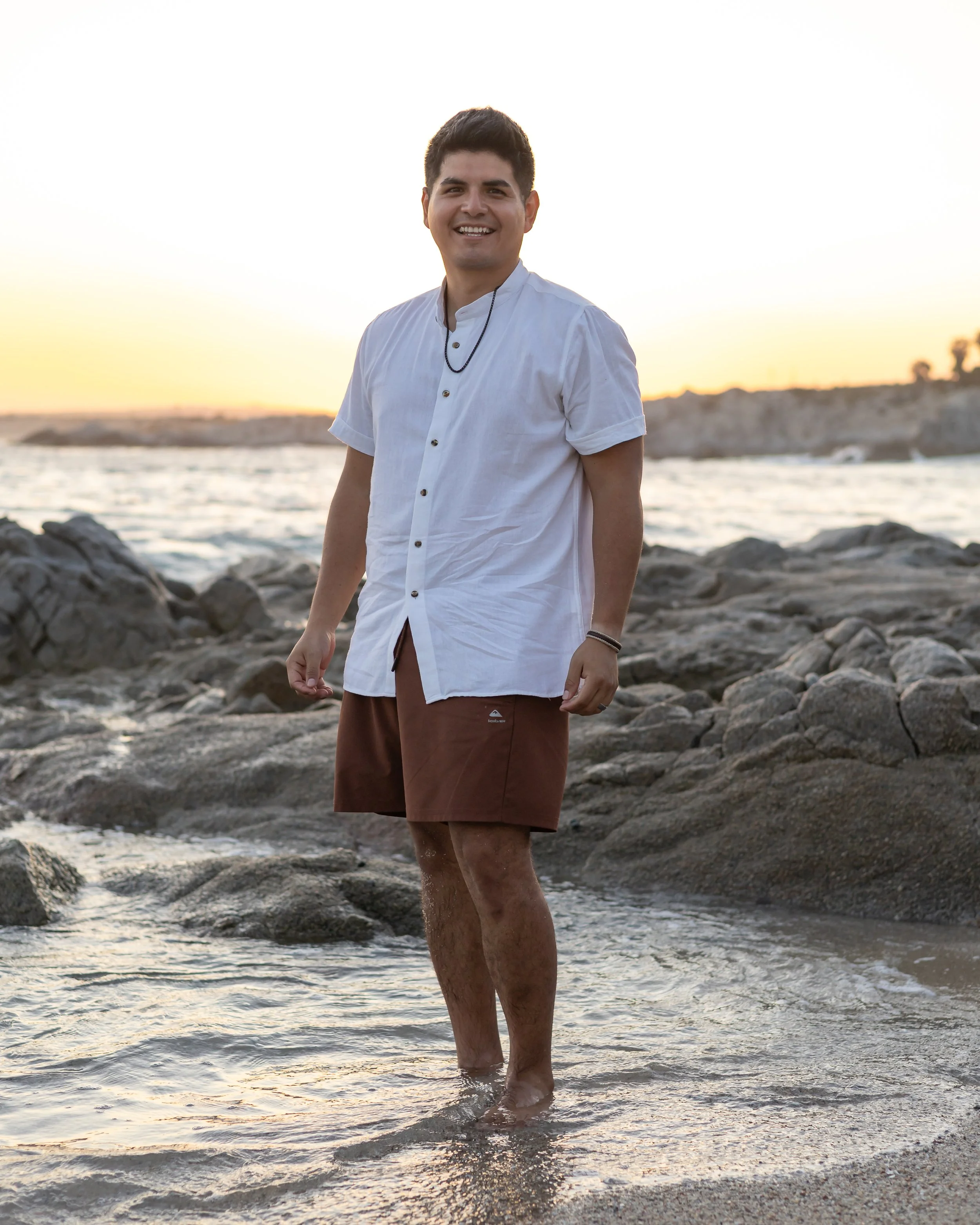 A young man is standing in shallow water at the beach during sunset. He has dark hair and is smiling, wearing a white short-sleeve button-up shirt, brown shorts, and a black necklace.