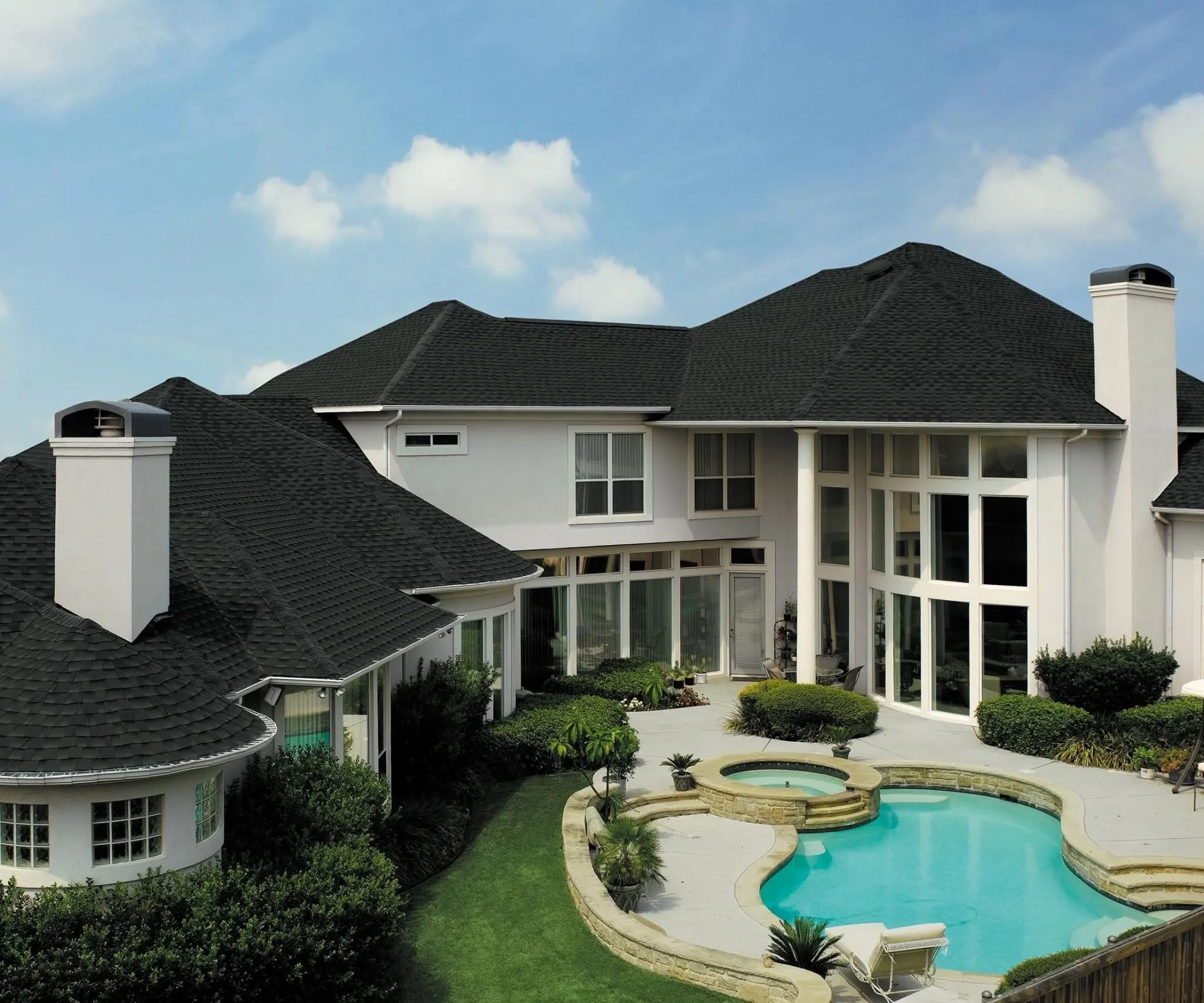 Large white house with black shingled roof and many windows, backyard with a freeform swimming pool, hot tub, and lush landscaping.