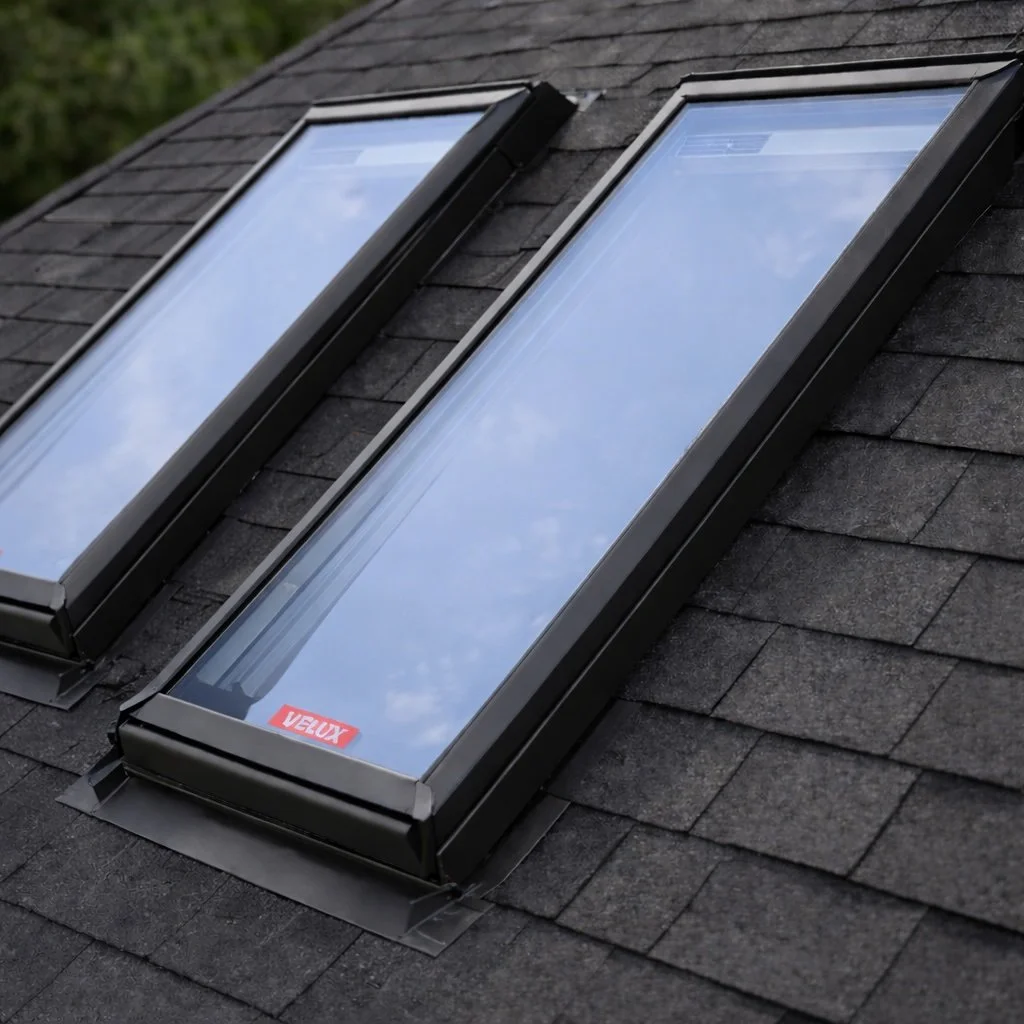 Two skylights installed on a black shingled roof, reflecting the sky and clouds.