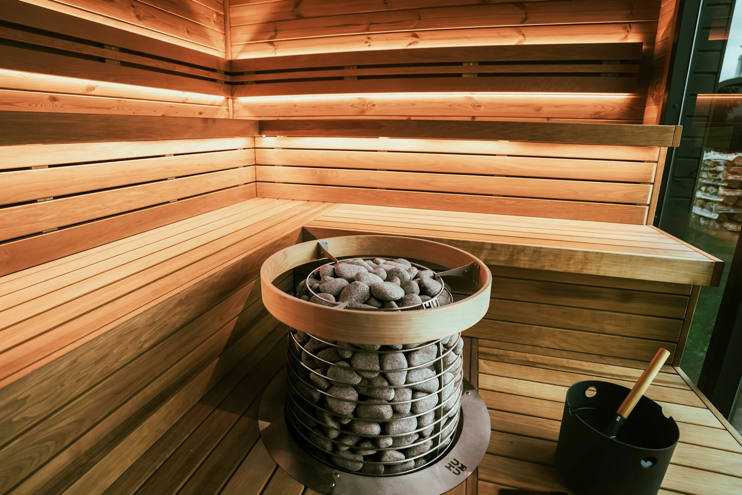 Wooden sauna interior with bench seating, a heater filled with stones, and a bucket with a ladle.
