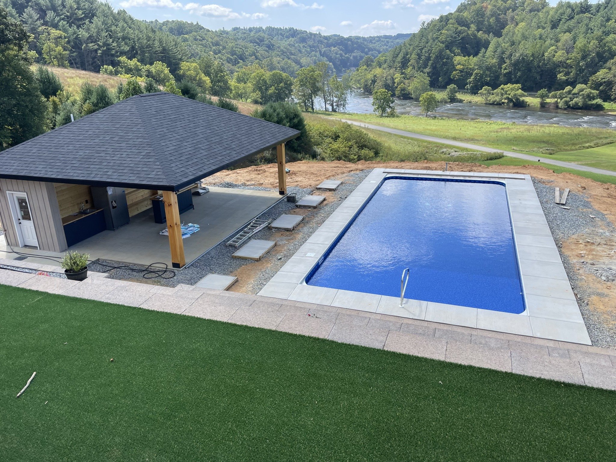 A backyard scene with a swimming pool, a covered outdoor structure, and a river in the background. The pool has a blue interior and is surrounded by a paved area. The outdoor structure has a black roof supported by wooden posts, with some construction materials nearby. The green lawn and scenic view of trees and water are visible in the background.