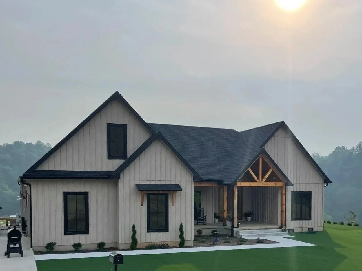 Modern farmhouse-style house with wood siding, black window frames, a front porch with steps, and a well-maintained lawn against a hilly background.
