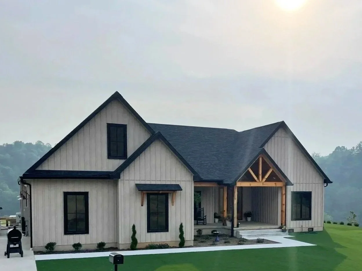 A new modern farmhouse with black window frames and a covered porch with wooden beams, situated on a grassy lawn with a mailbox in the foreground.