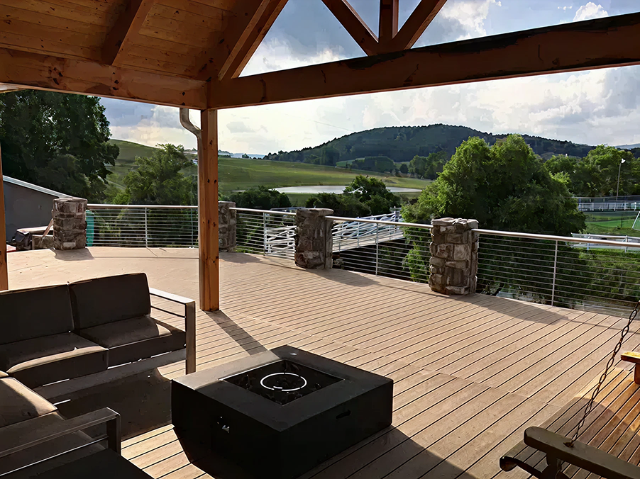 A spacious outdoor deck with a black fire pit, cushioned seating, a wooden swing, and a view of rolling hills, trees, and a partly cloudy sky.