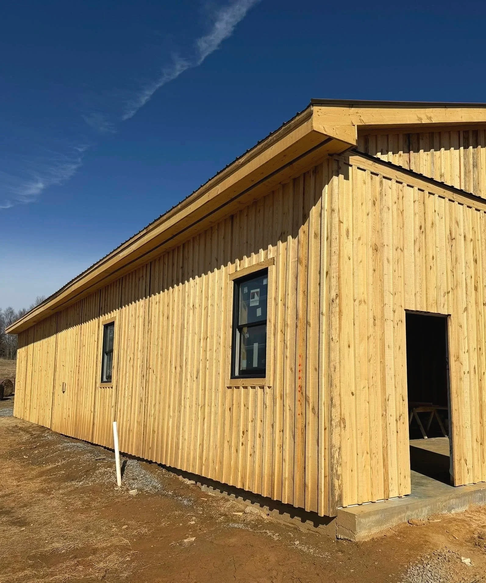 A partially constructed wooden house with vertical siding, two windows, and an open doorway under a clear blue sky.
