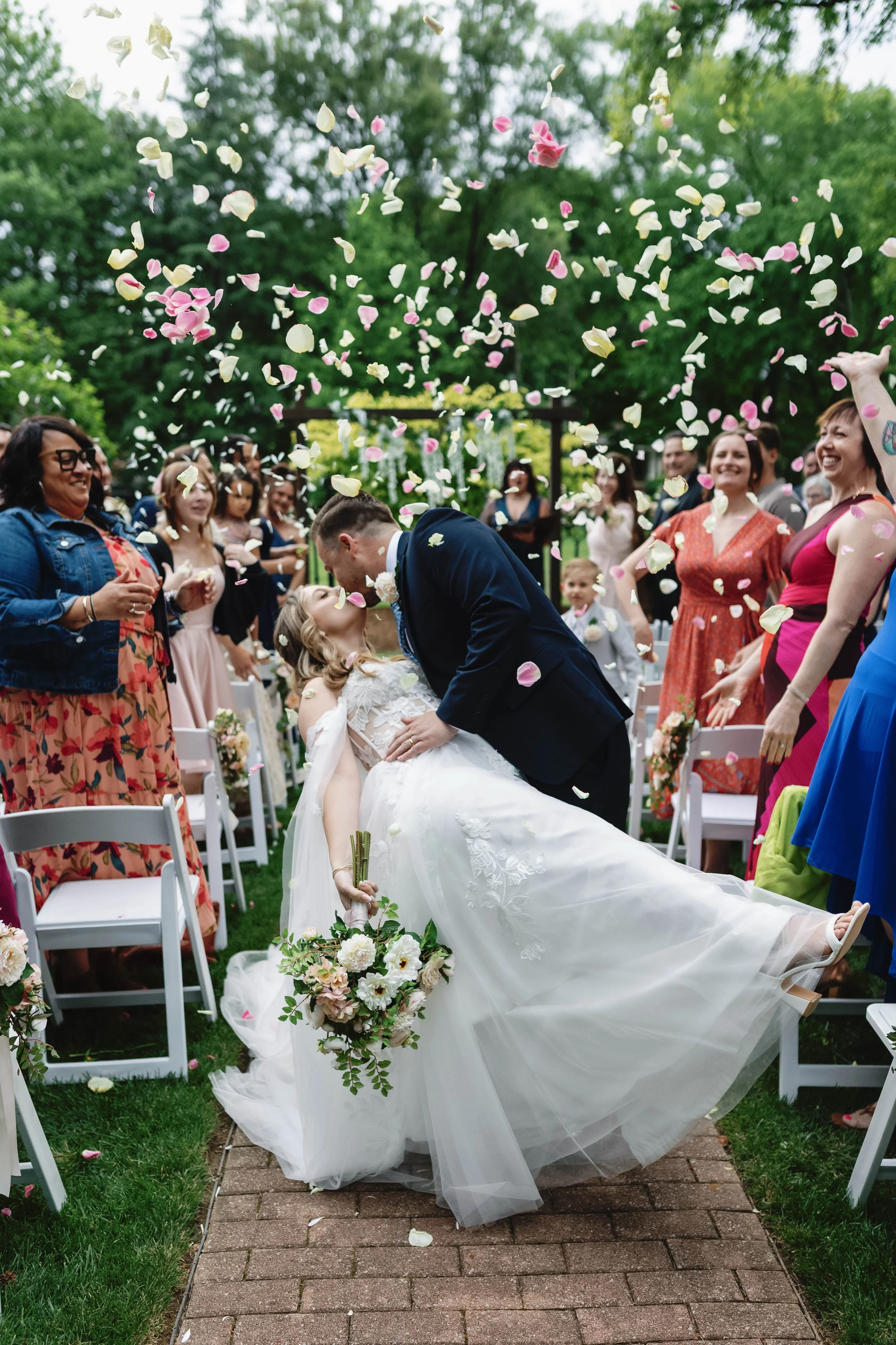 A newlywed couple kissing at their outdoor wedding ceremony, surrounded by friends and family, with flower petals falling from above. Peterson-Dumesnil house wedding kentucky