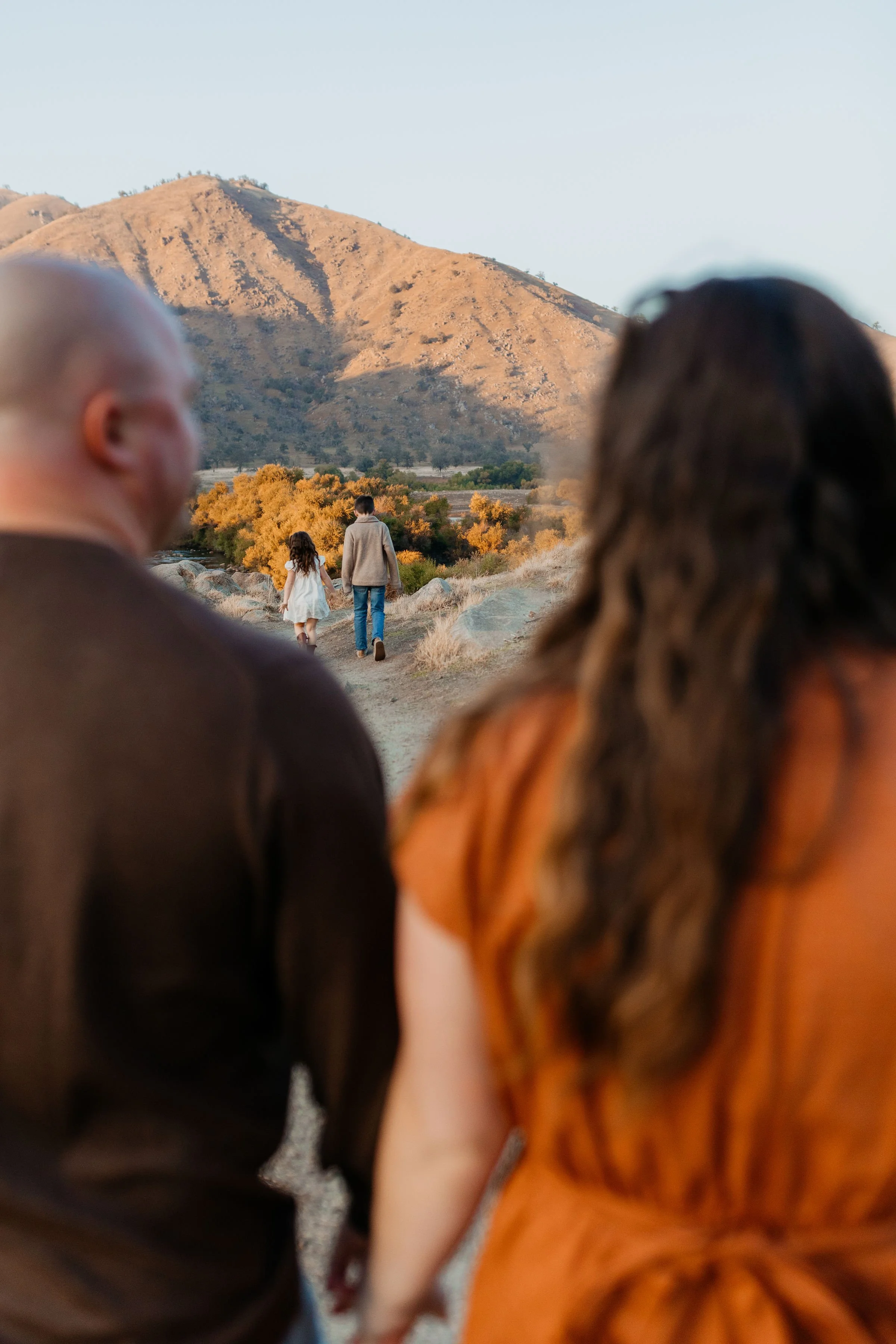 A couple holding hands and walking on a dirt trail, with children walking ahead towards a mountainous landscape with autumn-colored trees.  lexington kentucky family photographer storytelling portraits