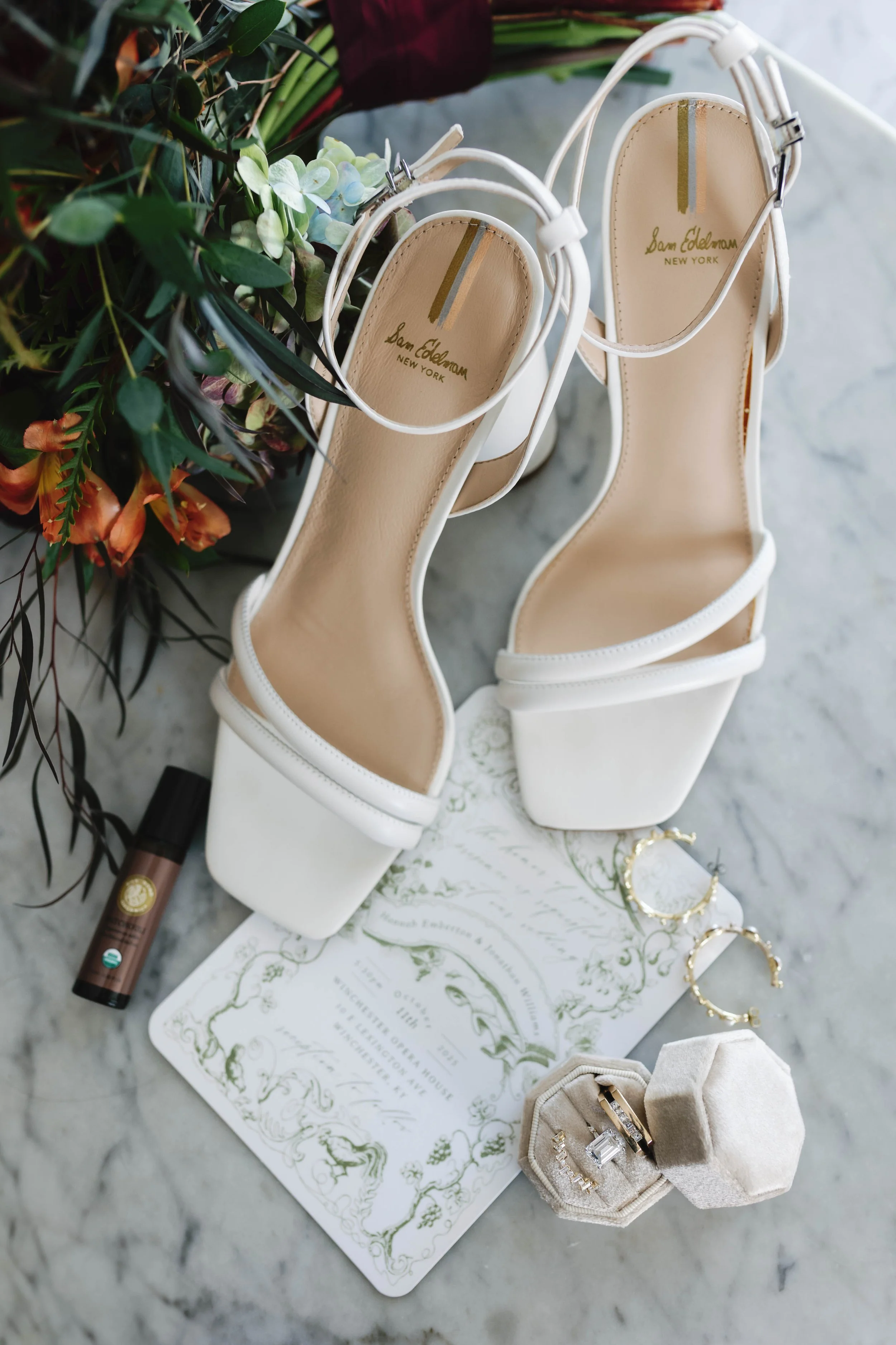White high-heeled sandals with ankle straps, gold hoop earrings, a jewelry box with rings, a perfume bottle, and a wedding invitation on a marble surface moundale manor historic kentucky wedding venue winchester kentucky 
