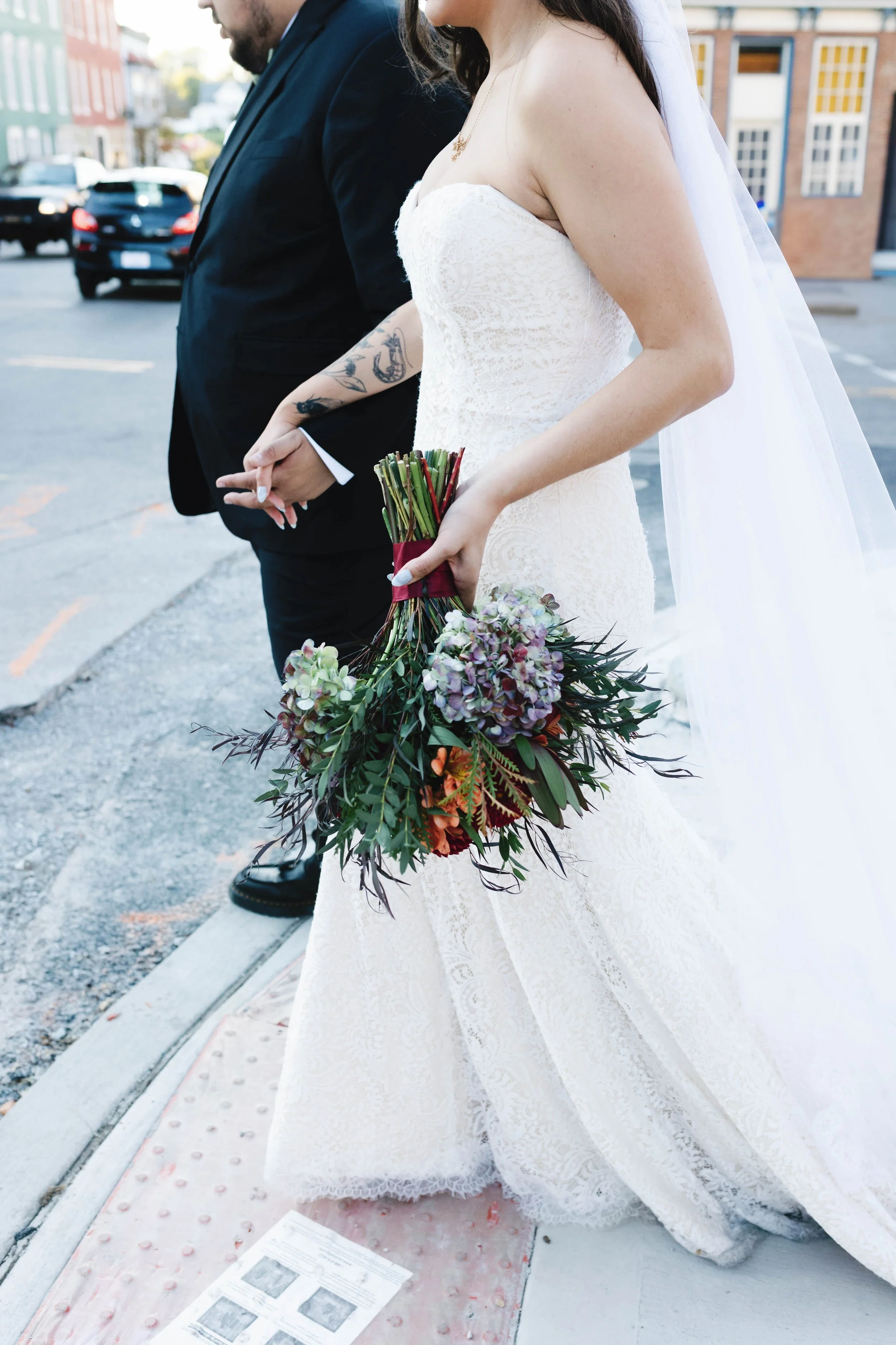 A bride in a white lace wedding dress holding a bouquet of flowers, walking with a groom in a dark suit and tie, outside on a city street. winchester opera house historic wedding kentucky 