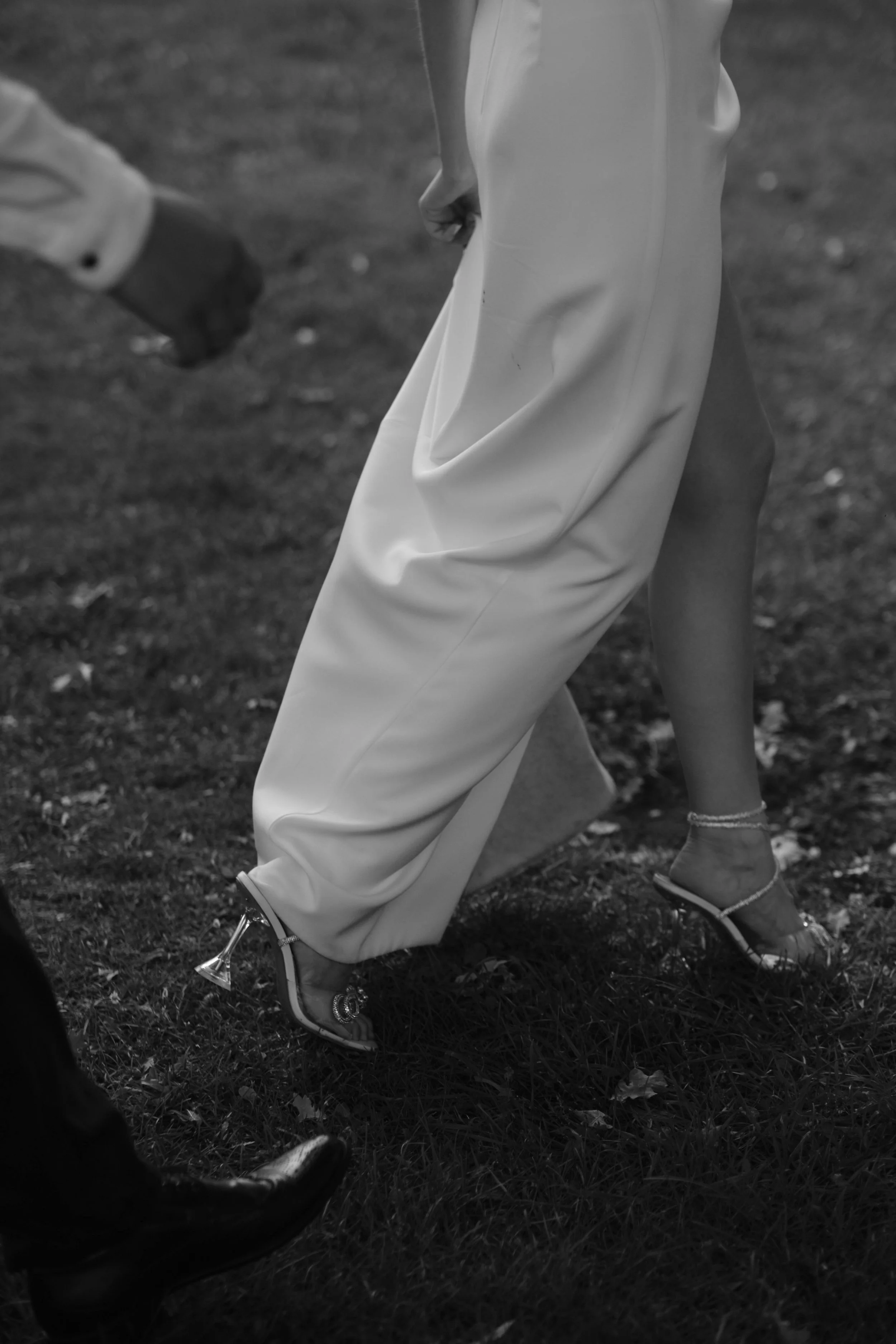 Close-up of a person wearing elegant high-heeled shoes with thin ankle straps and a flowing dress, standing on grass, with another person's leg visible on the left.
