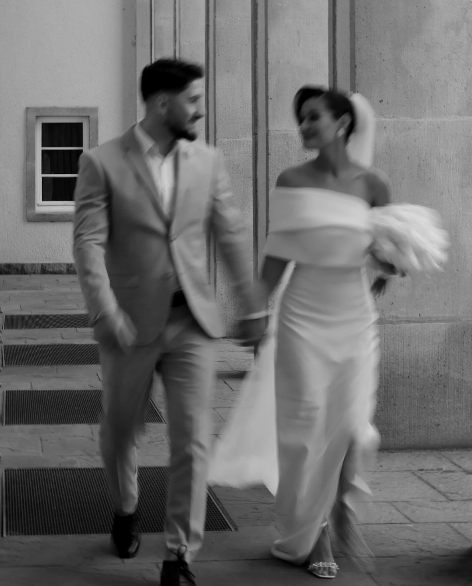 A man and woman in formal attire walking together outside at night, holding hands, with a stone wall behind them.