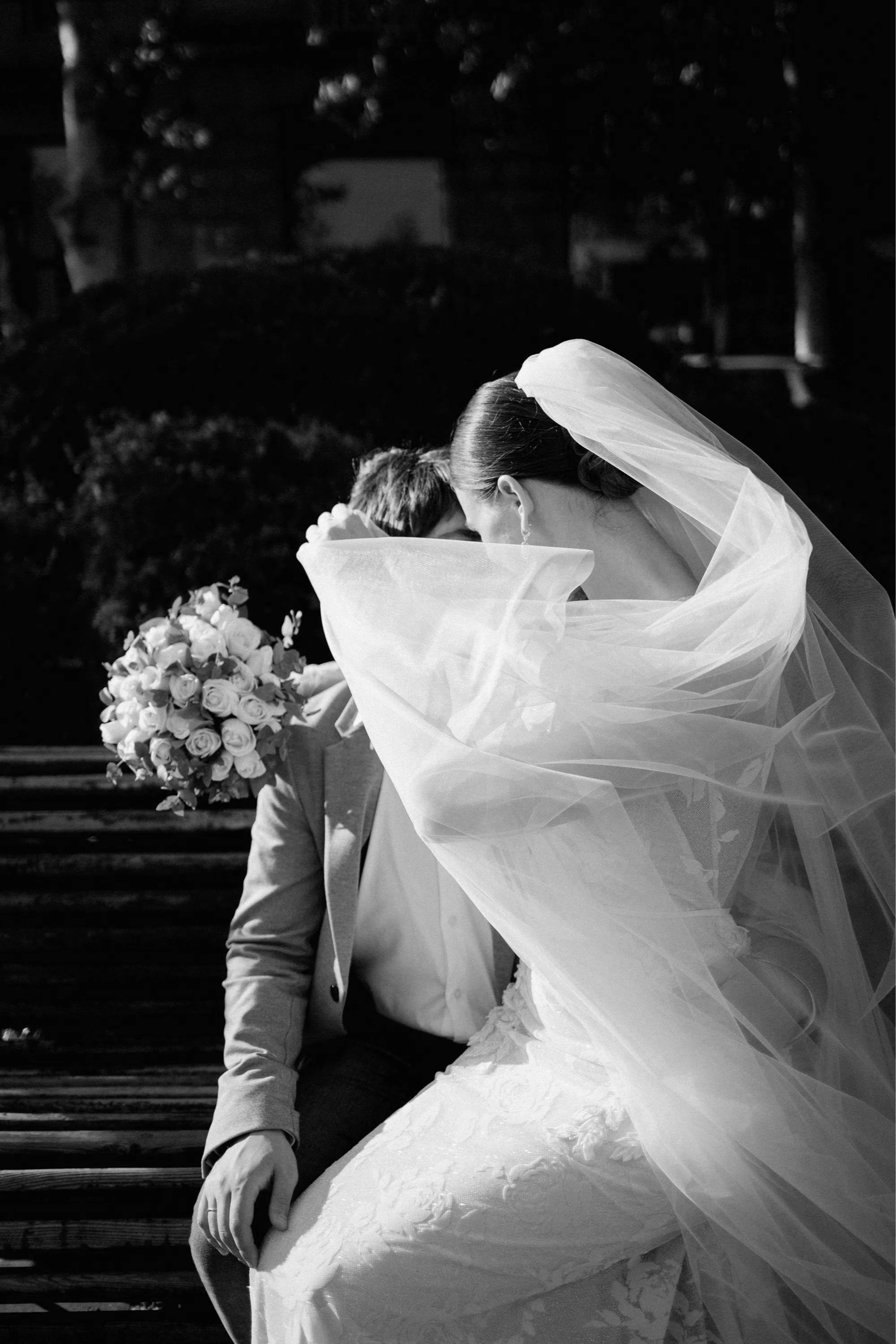 A bride and groom sit on a park bench, sharing a kiss under the bride's veil during their wedding day. The bride holds a bouquet of roses, and the scene is captured in black and white.