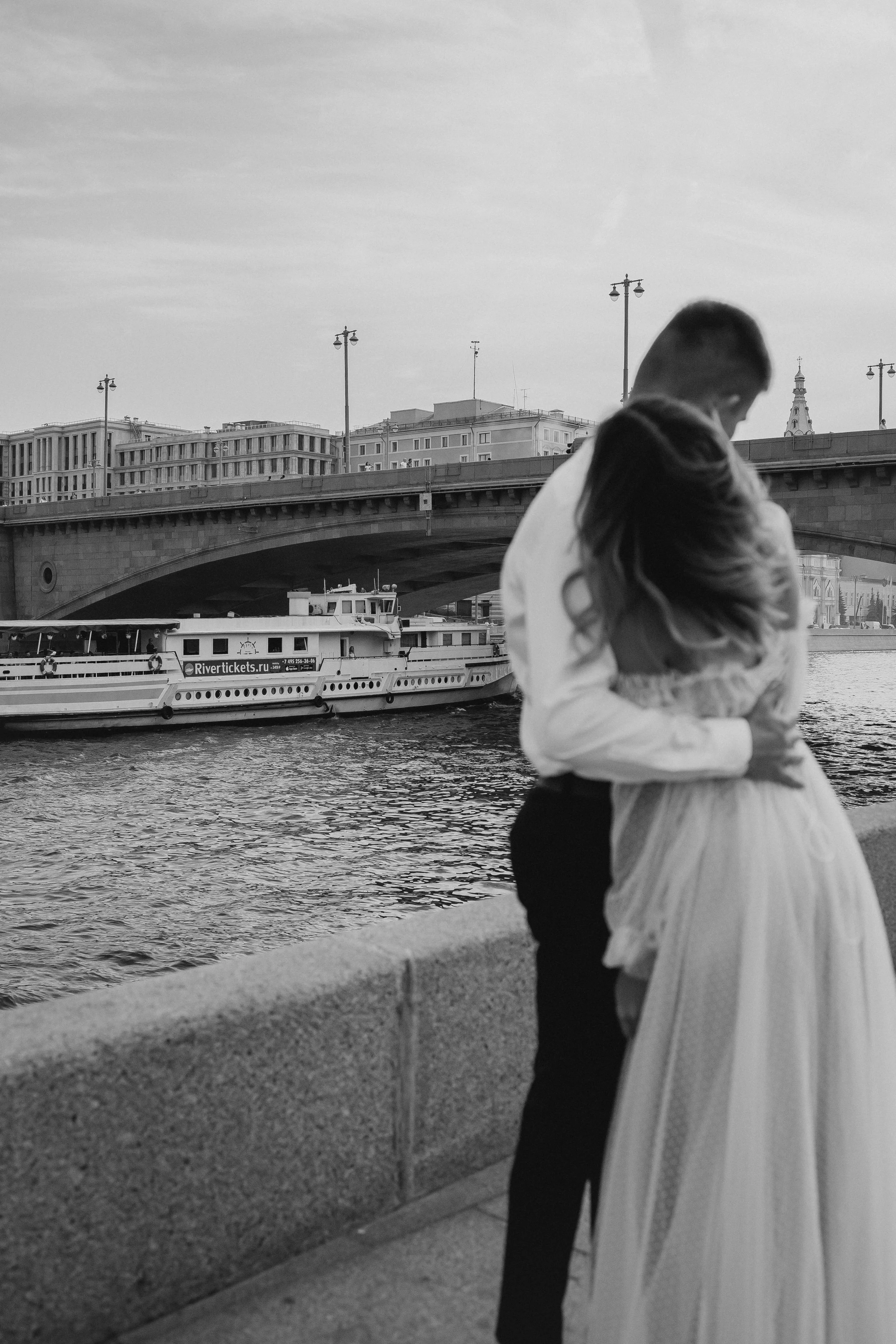 A couple embracing near a river with a boat and bridge in the background, black and white photo.