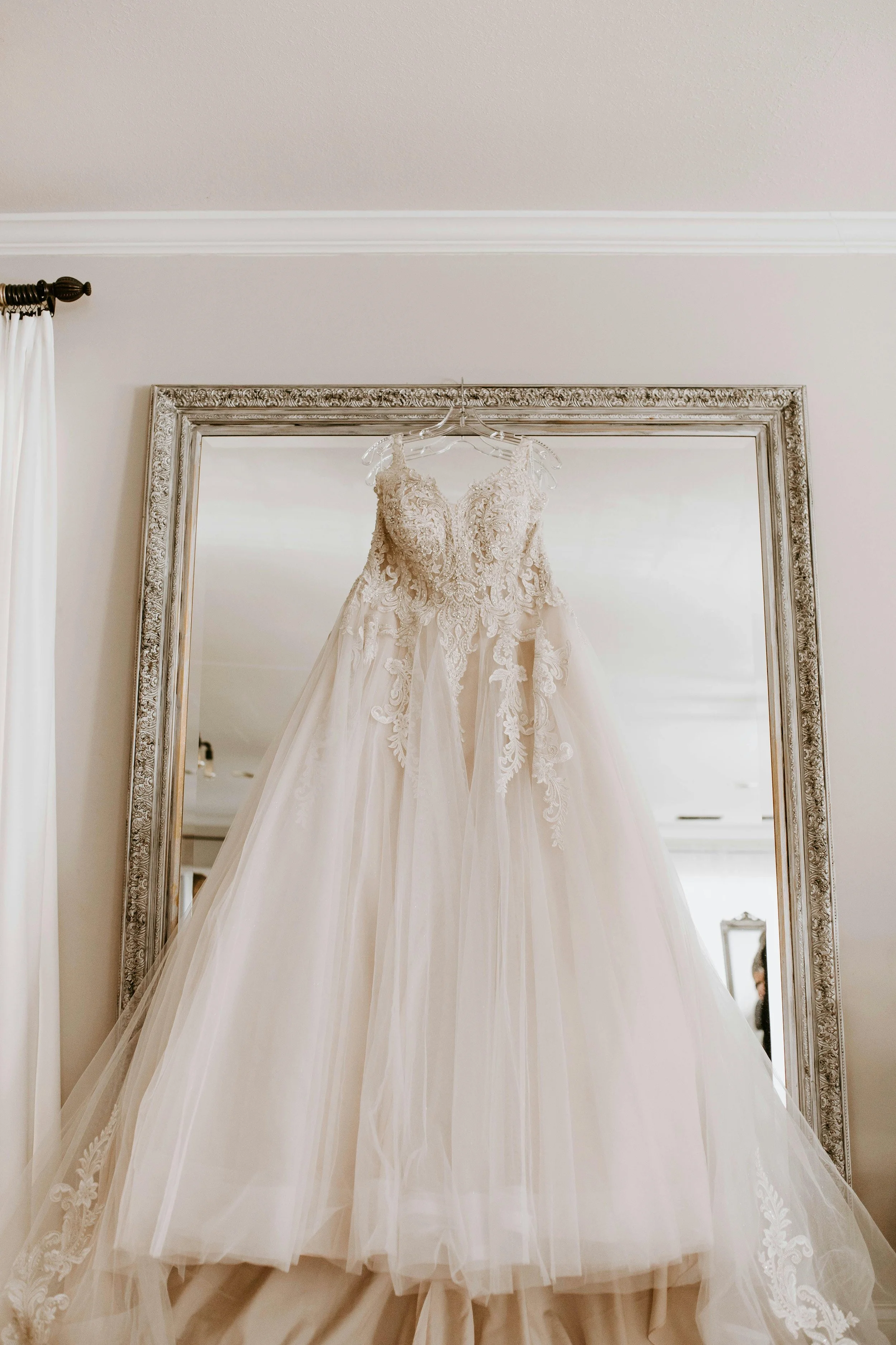 Elegant wedding gown with lace details hanging in front of a large mirror.