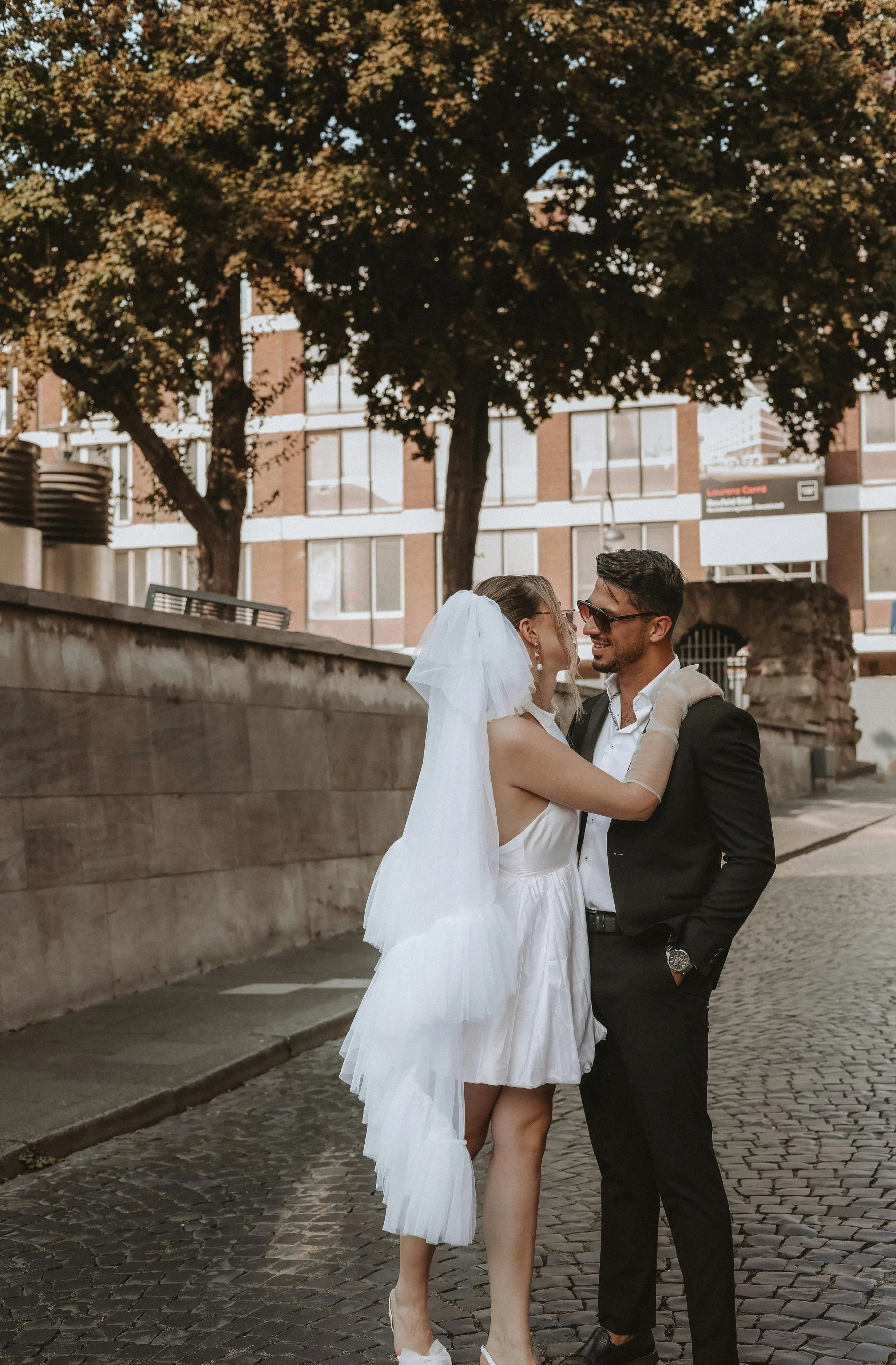 A bride and groom smiling at each other on a cobblestone street in an urban area, surrounded by trees and buildings.