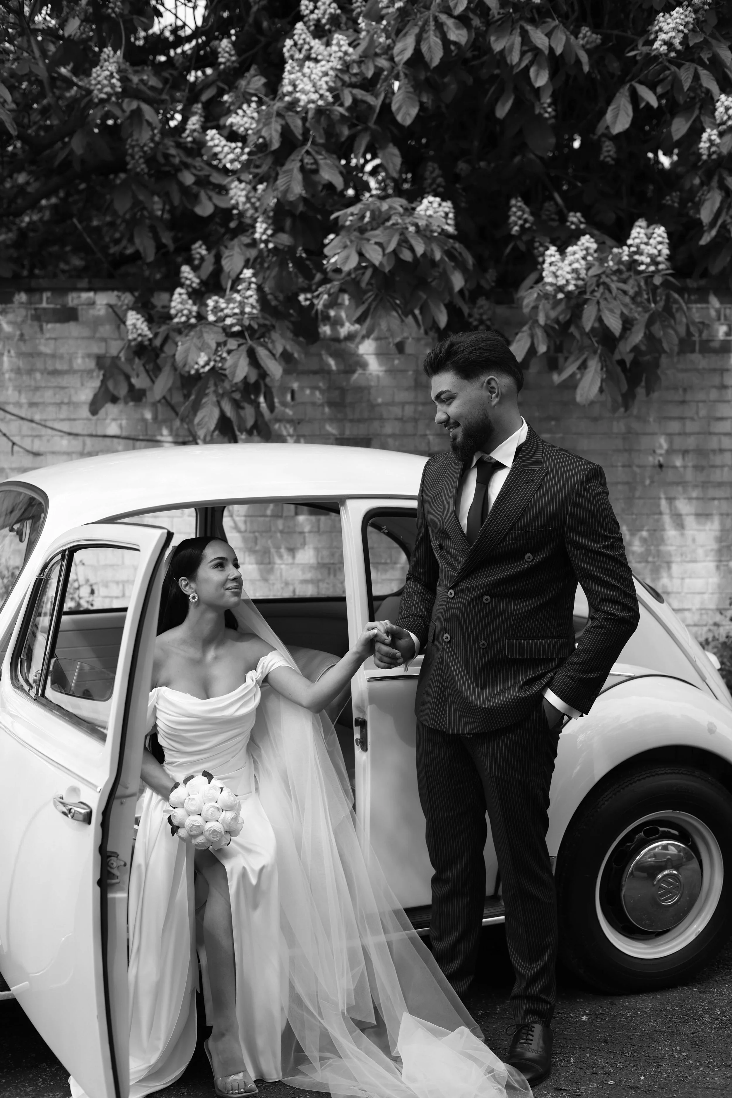 Black and white photo of a bride sitting inside a vintage car, holding a bouquet of roses, and a groom standing outside the car, holding her hand, both smiling. The background features a brick wall and leafy trees.