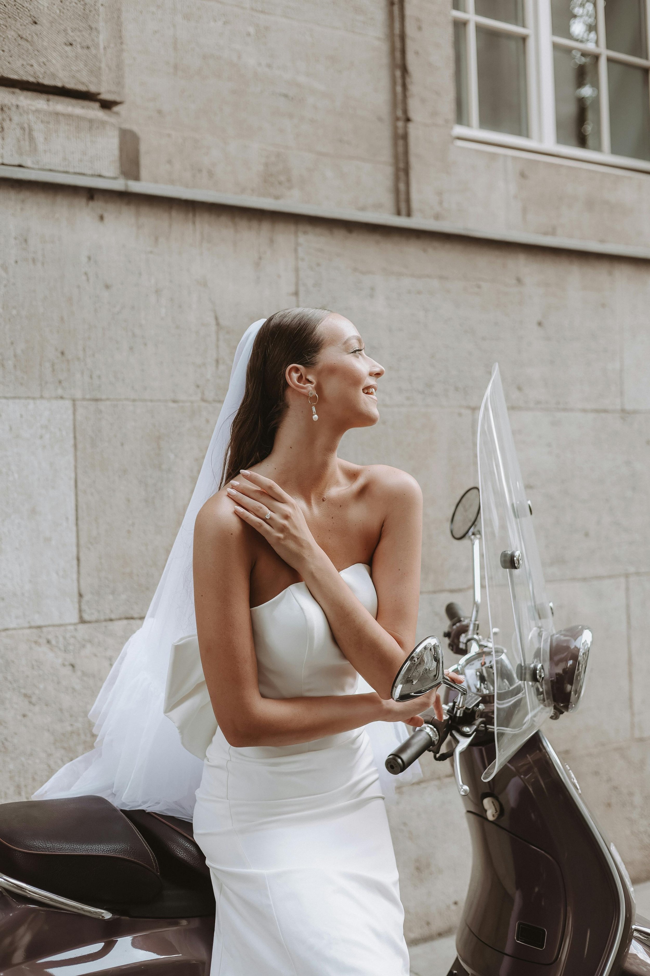 A smiling bride with long brown hair, wearing a strapless white wedding dress and a veil, sitting on a motorcycle with a windscreen, against a stone building wall.