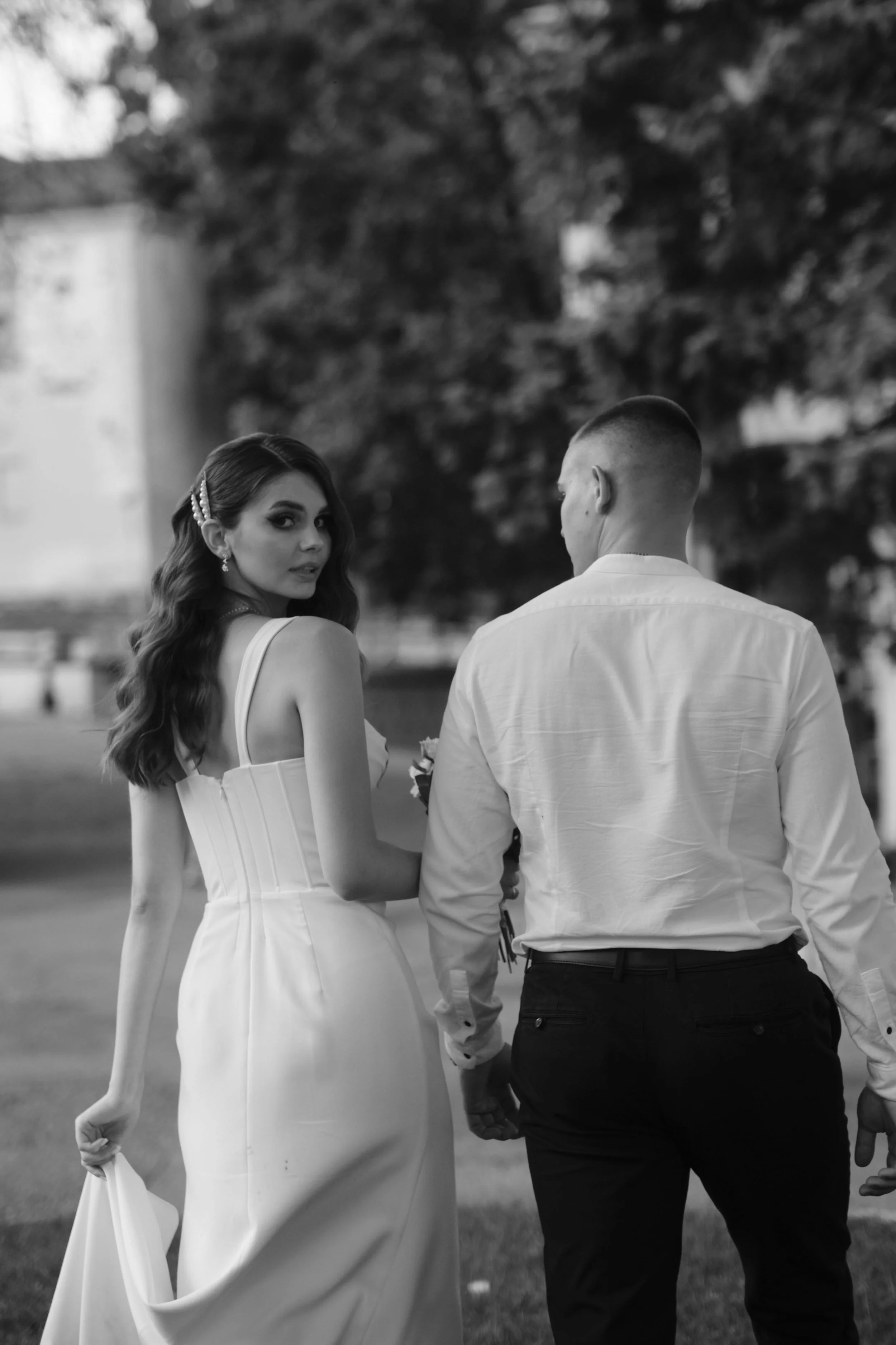 A black-and-white photograph of a couple holding hands outdoors, with the woman looking back over her shoulder. The woman is wearing a strapless white dress, and the man is dressed in a white shirt and dark pants. Background includes trees and blurre