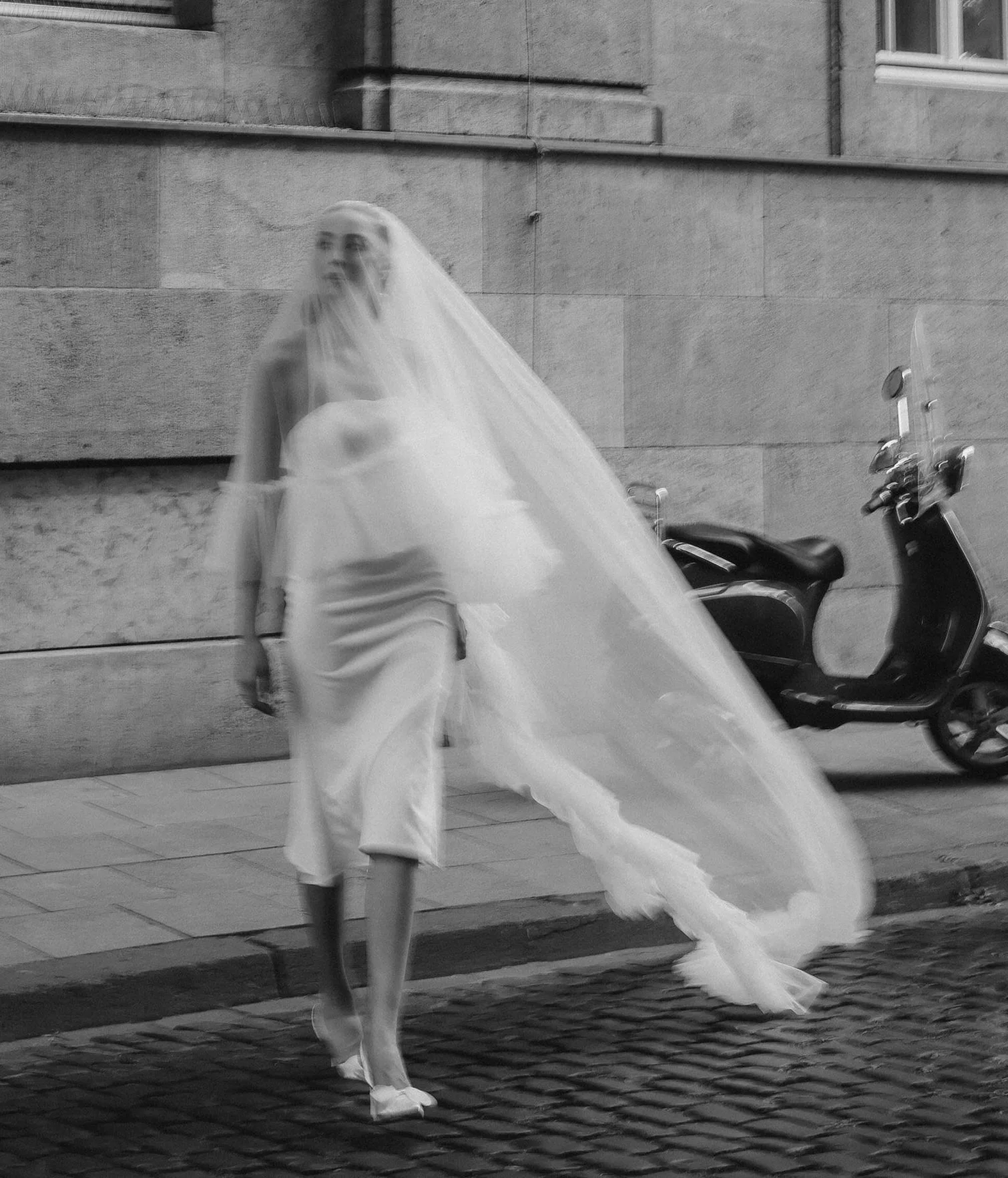 A woman in a white dress and veil walking on a city street beside a scooter and stone building wall.