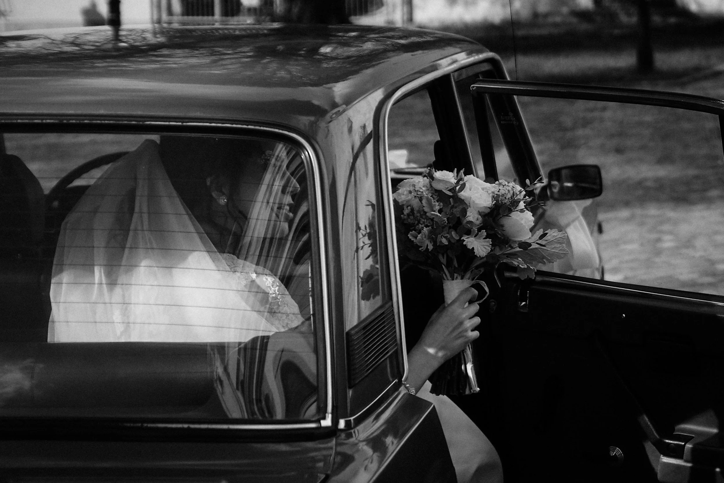 A bride sitting inside a vintage car, holding a bouquet of flowers through the open passenger door.