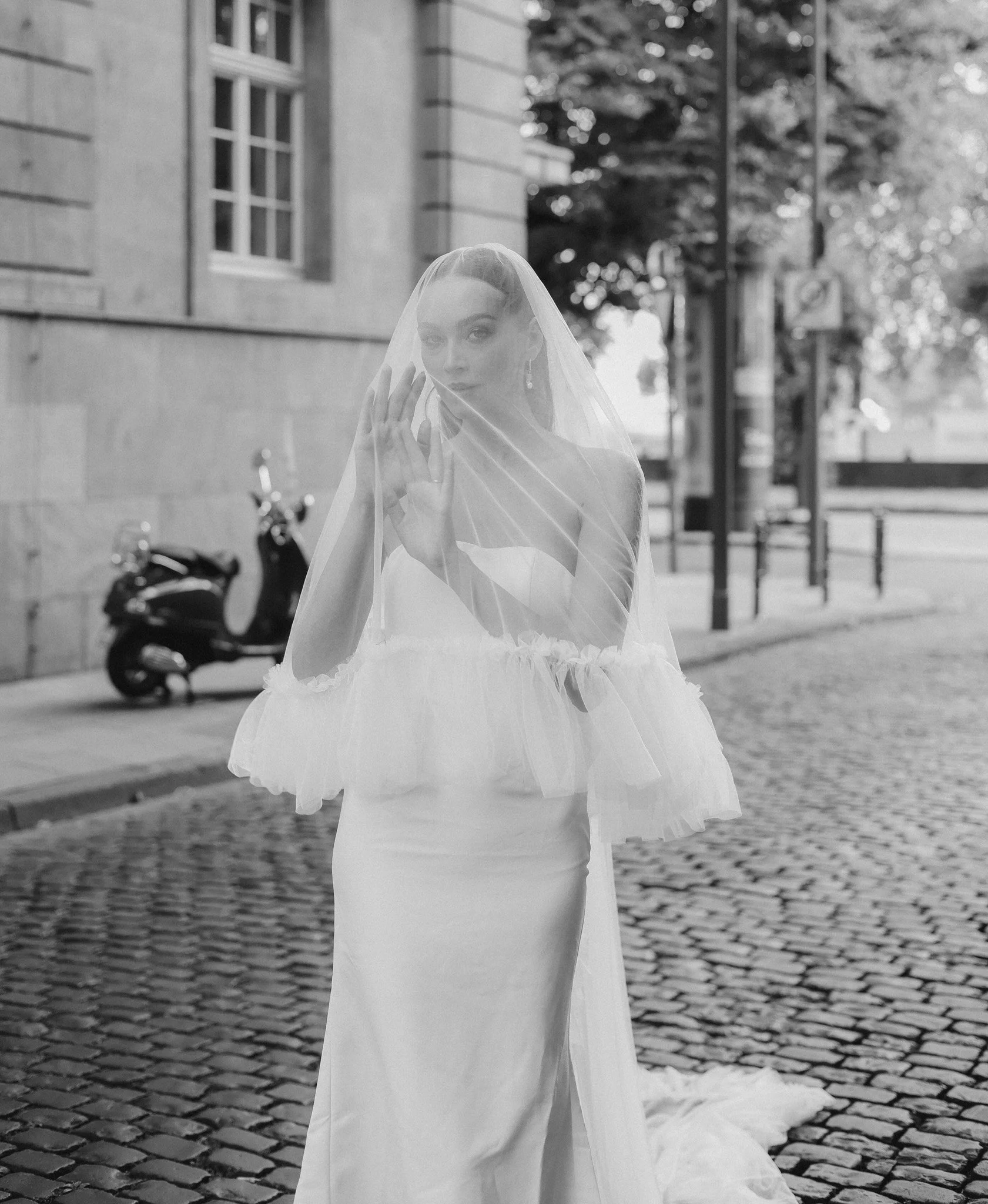 A woman wearing a wedding dress and veil stands on a cobblestone street, looking at the camera with her hand near her face, with buildings and a scooter in the background.