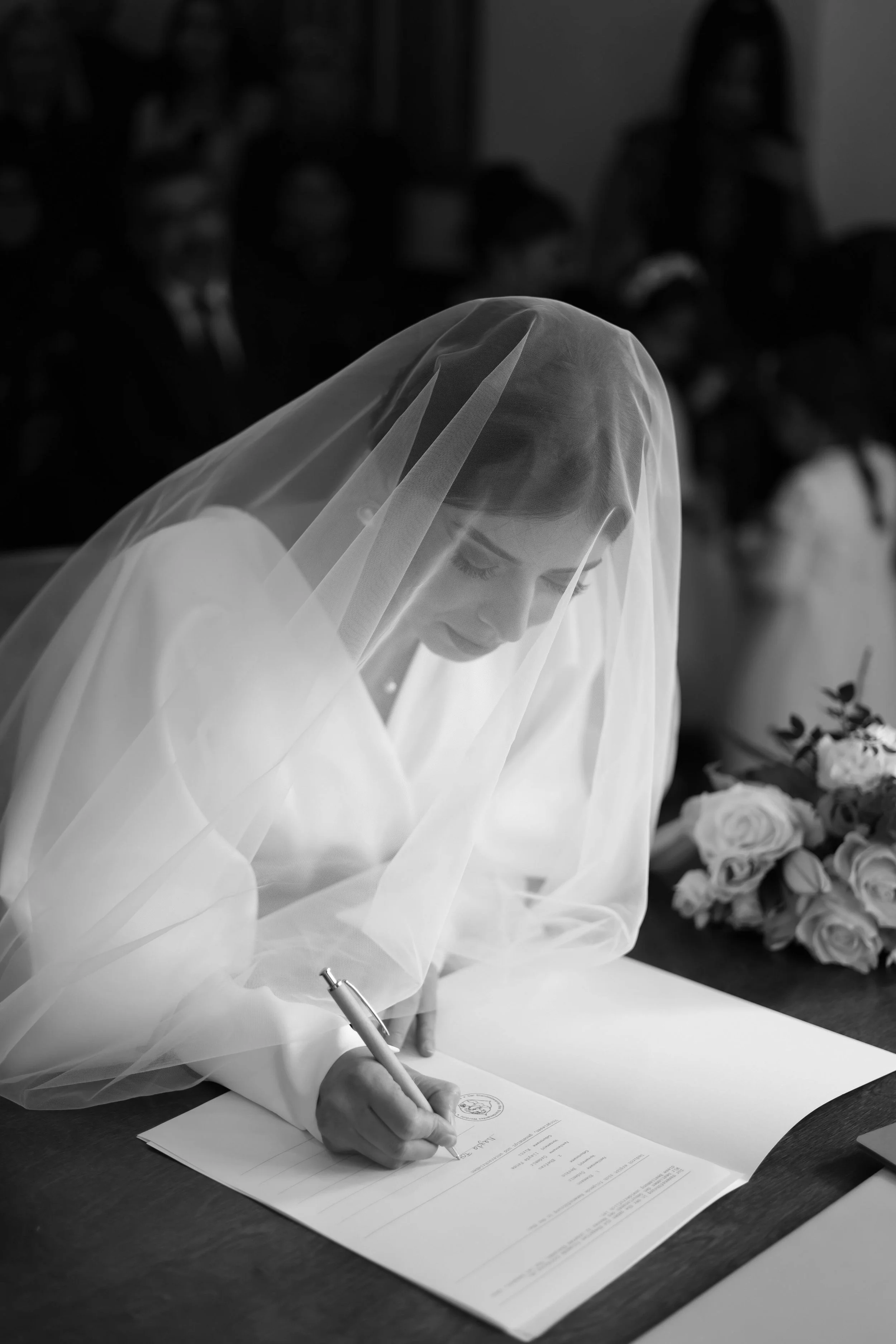 A bride in a wedding gown and veil signing a document at her wedding.