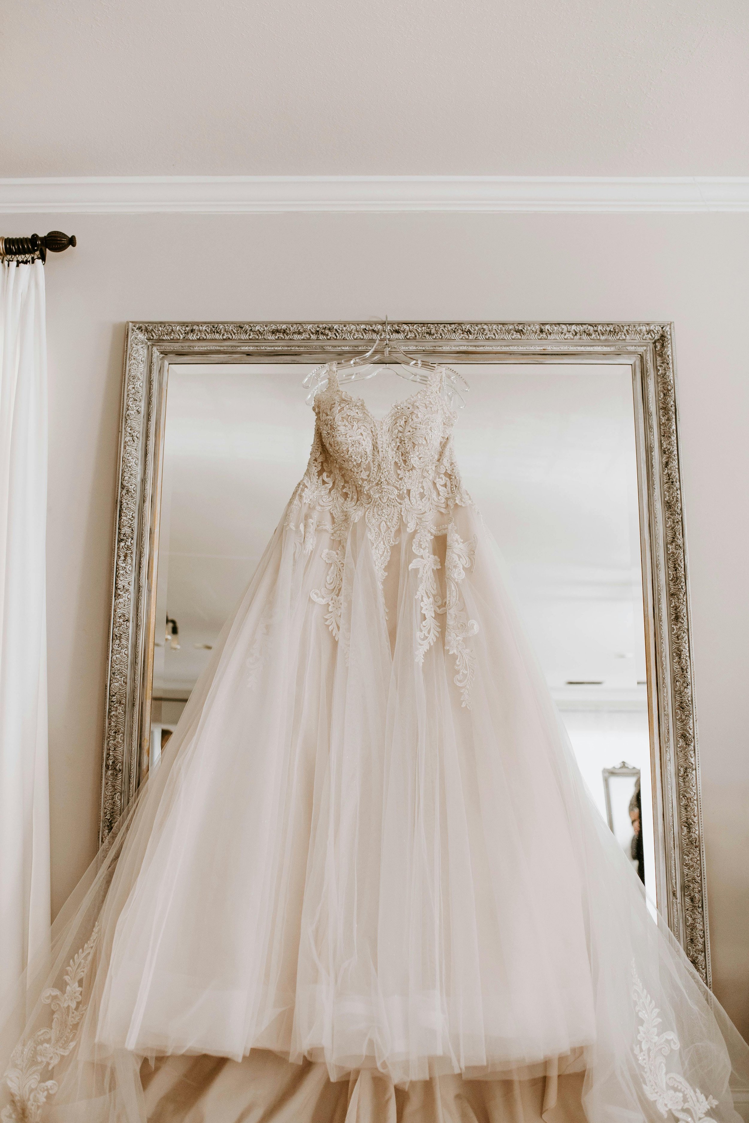 A wedding dress with intricate lace details hanging on a hanger in front of a large mirror.
