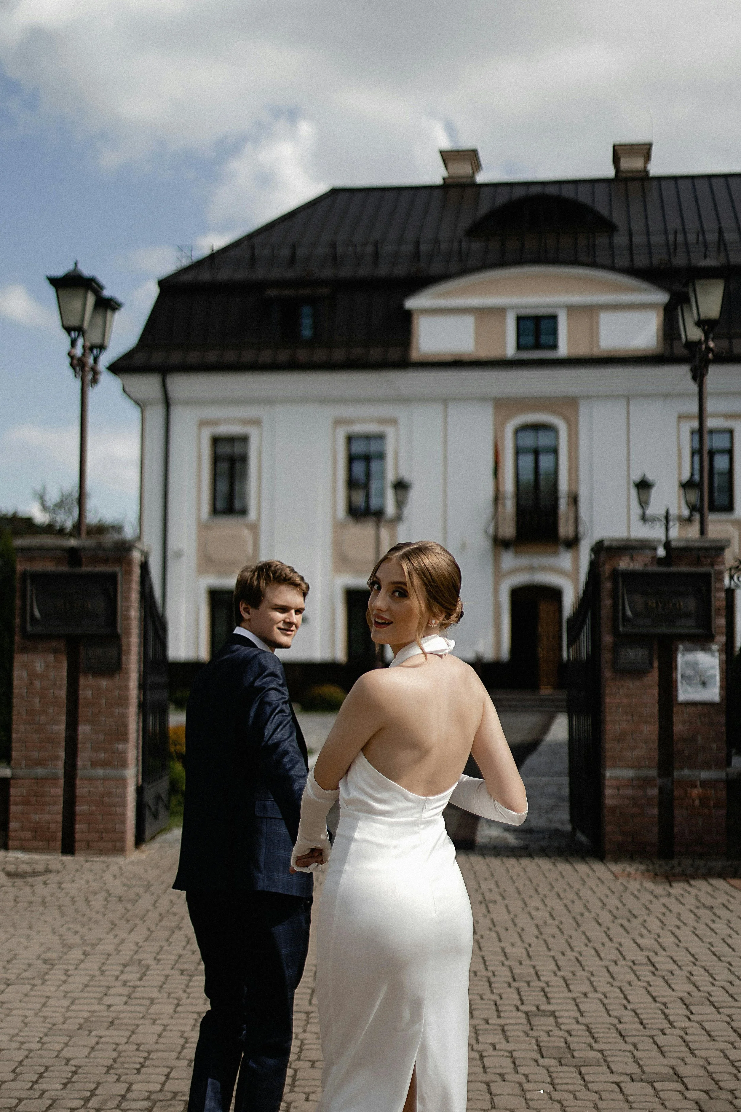 A woman in a white wedding dress and gloves stands in front of a large white house with a dark roof, turning to smile at the camera while holding hands with a man in a dark suit, who is also smiling at the camera.