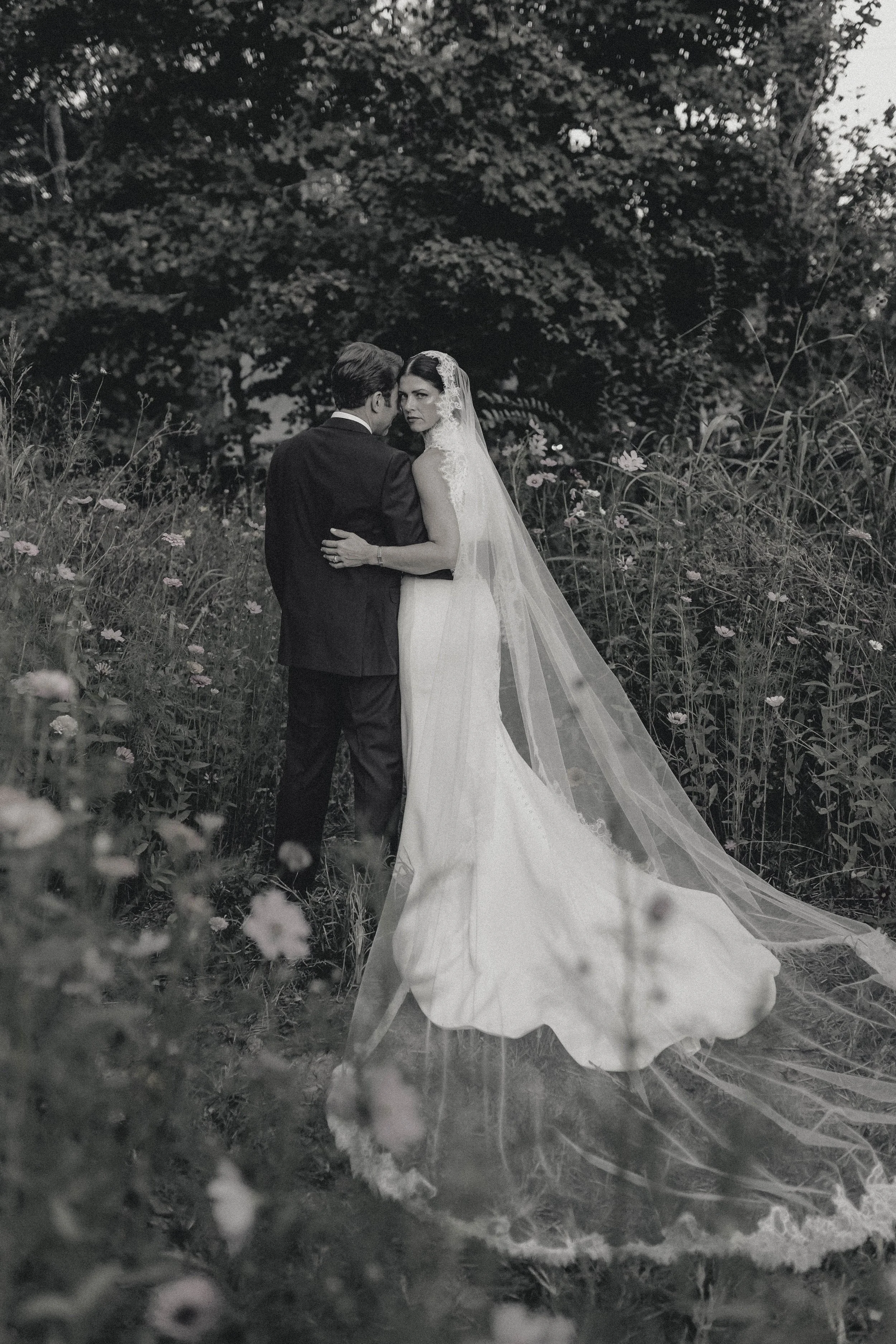 Black and white photo of a bride and groom standing close together in a natural outdoor setting with tall grass and flowers, lush trees in the background. elegant backyard kentucky wedding