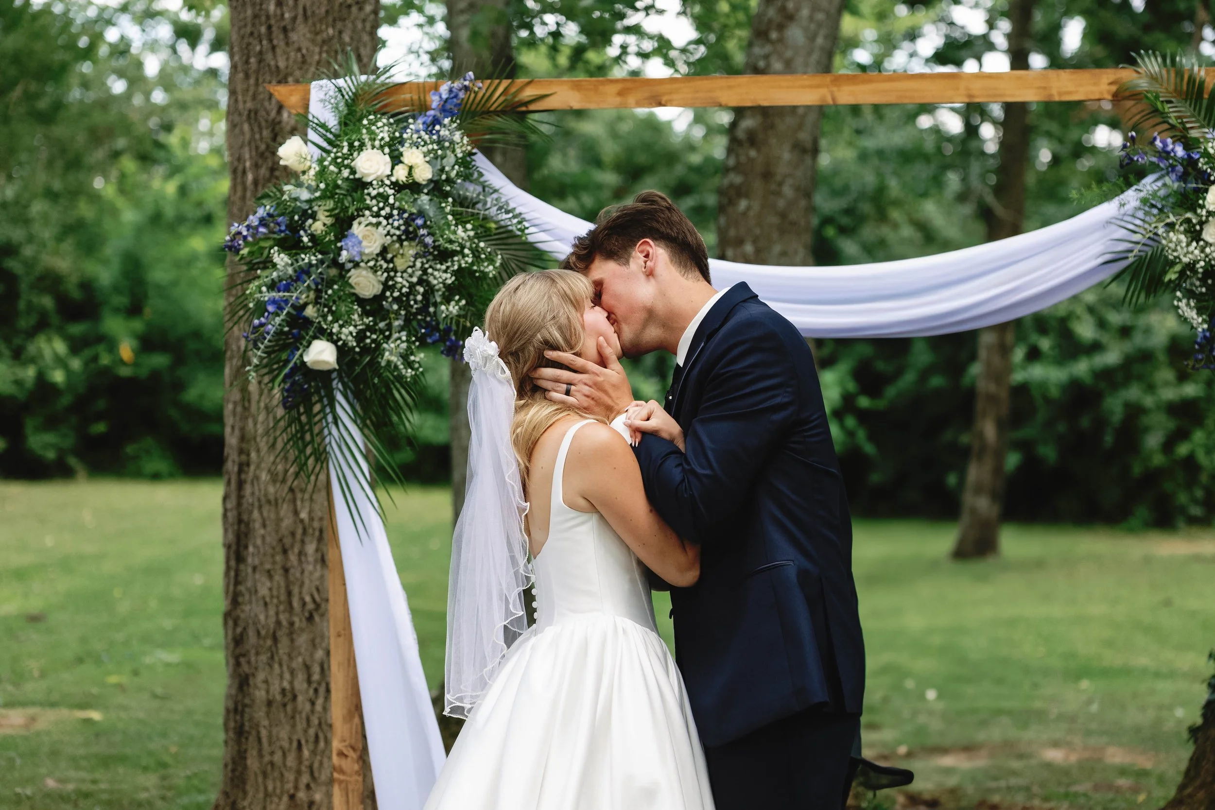 A bride and groom share a kiss beneath a wedding arch decorated with flowers and white draping outdoors with trees in the background. ashford acres inn wedding cynthiana kentucky
