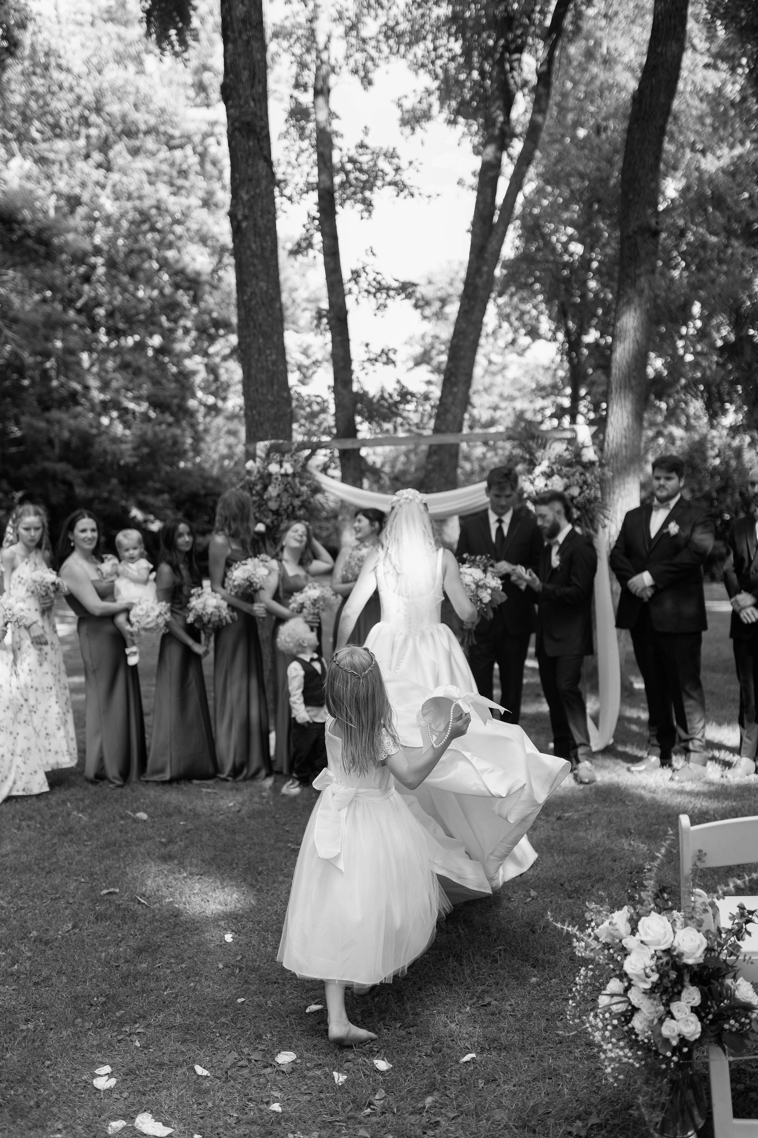 A wedding ceremony outdoors with the bride, bridesmaids, groomsmen, and guests gathered under tall trees, with a young girl running in front of the bride holding a ribbon or decoration.ashford acres inn wedding